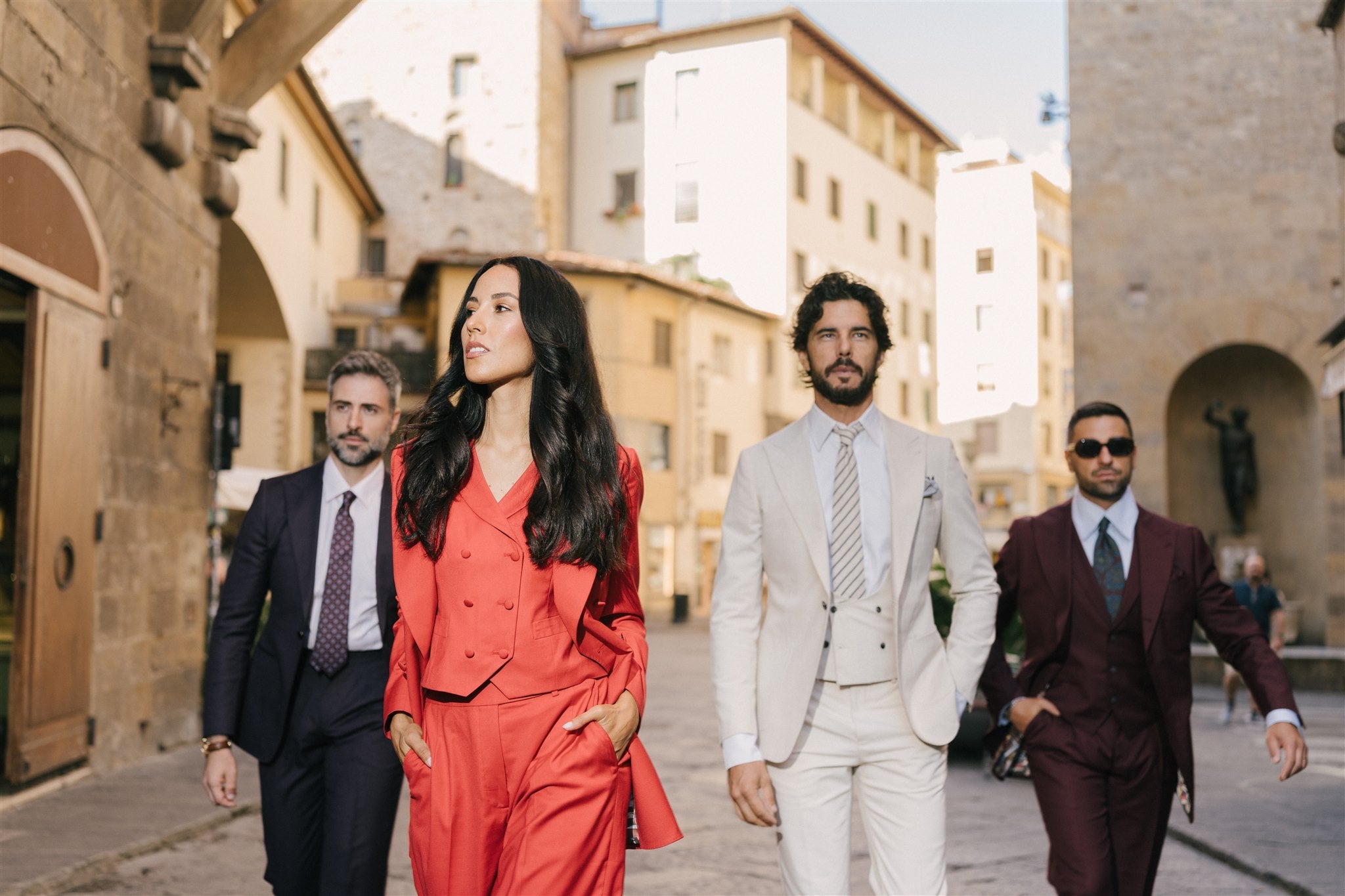 The image shows four fashionably dressed individuals confidently walking in a historic Florence street. Outfits feature vibrant and classic colors: red, ivory, burgundy, and dark navy suits, reflecting elegant Italian-inspired style.