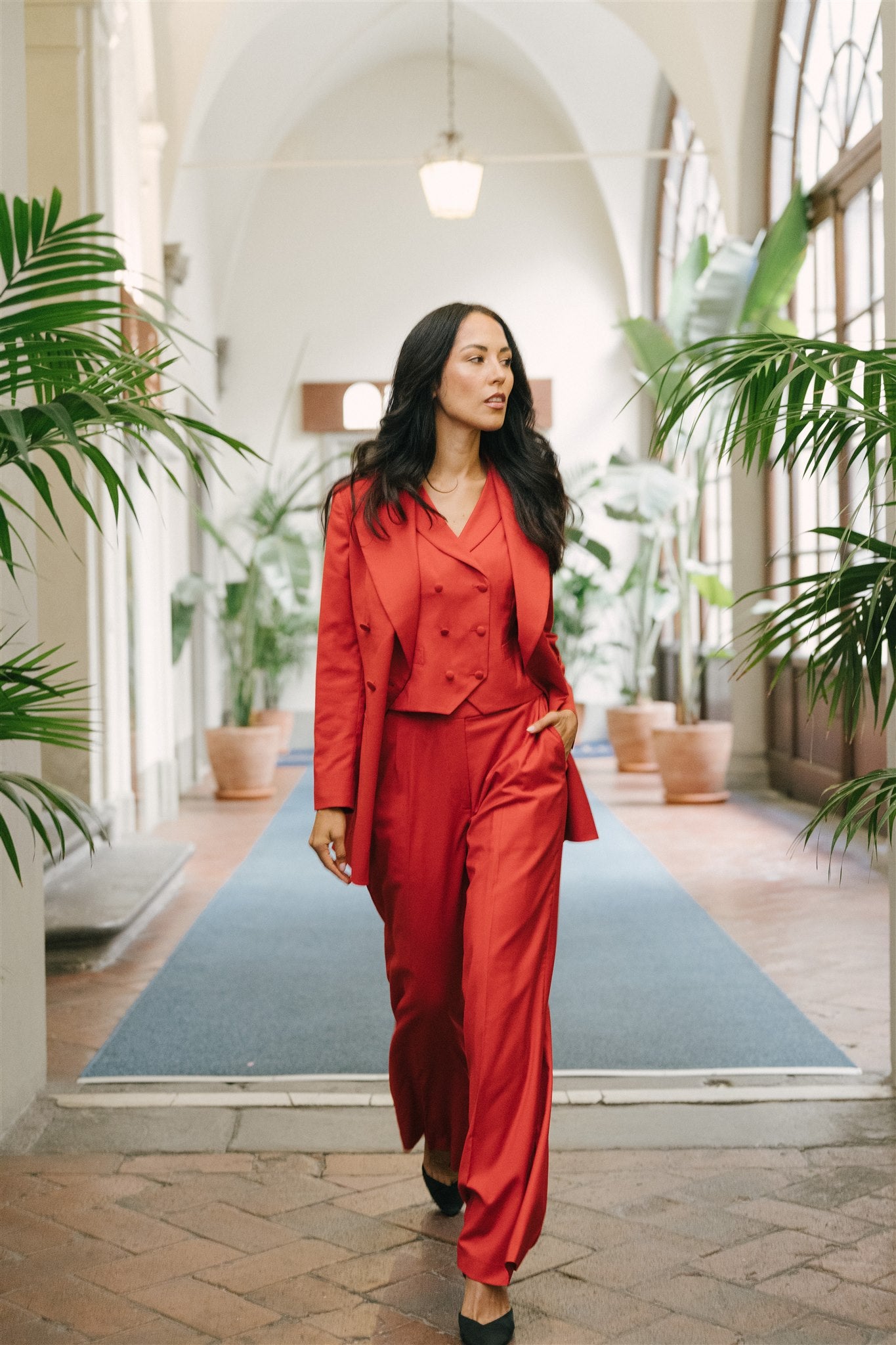 Woman in a red suit walking through an indoor setting with plants and arches.