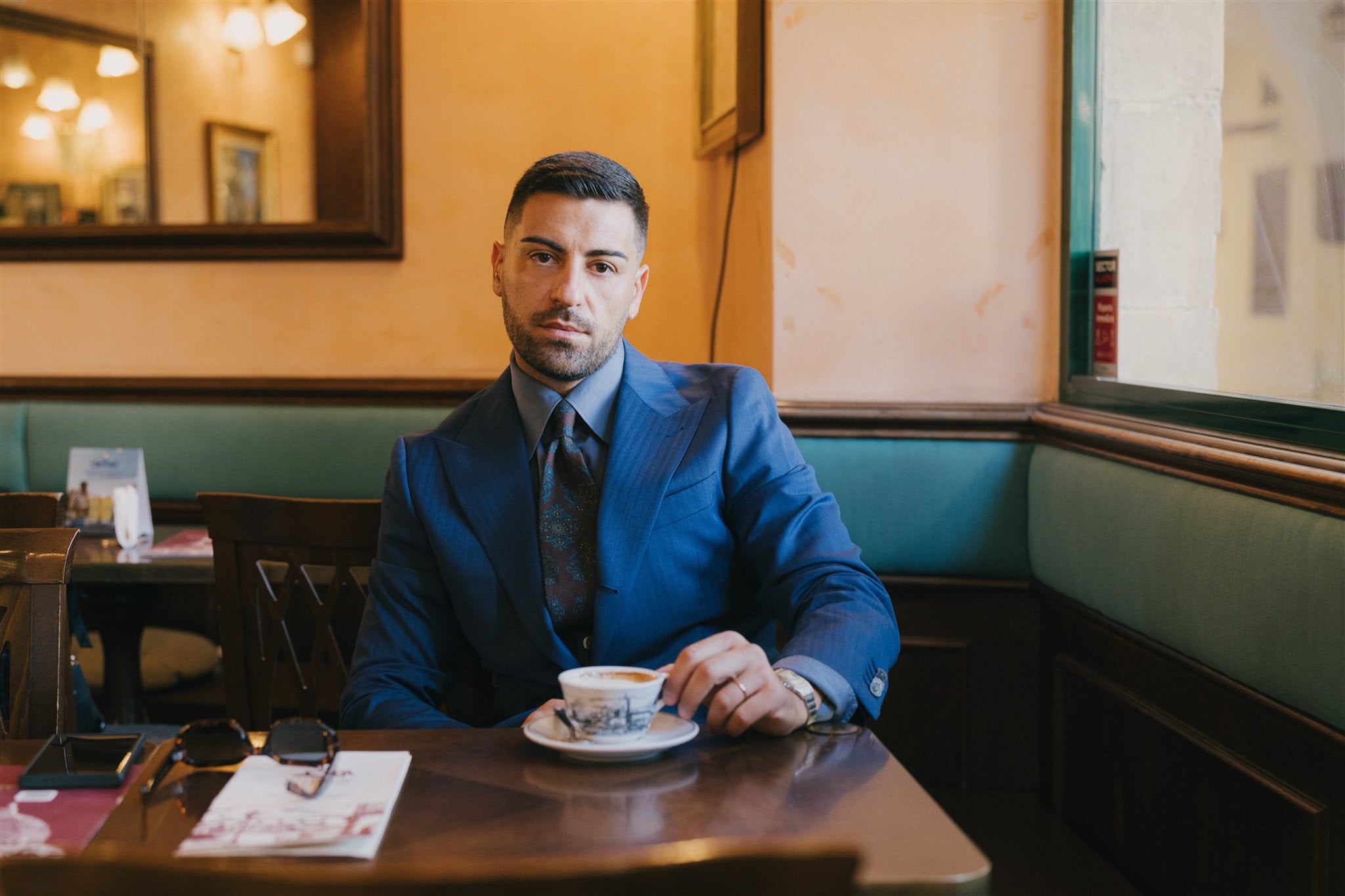 Man in a blue suit sitting at a table in a cafe with a cup of coffee.