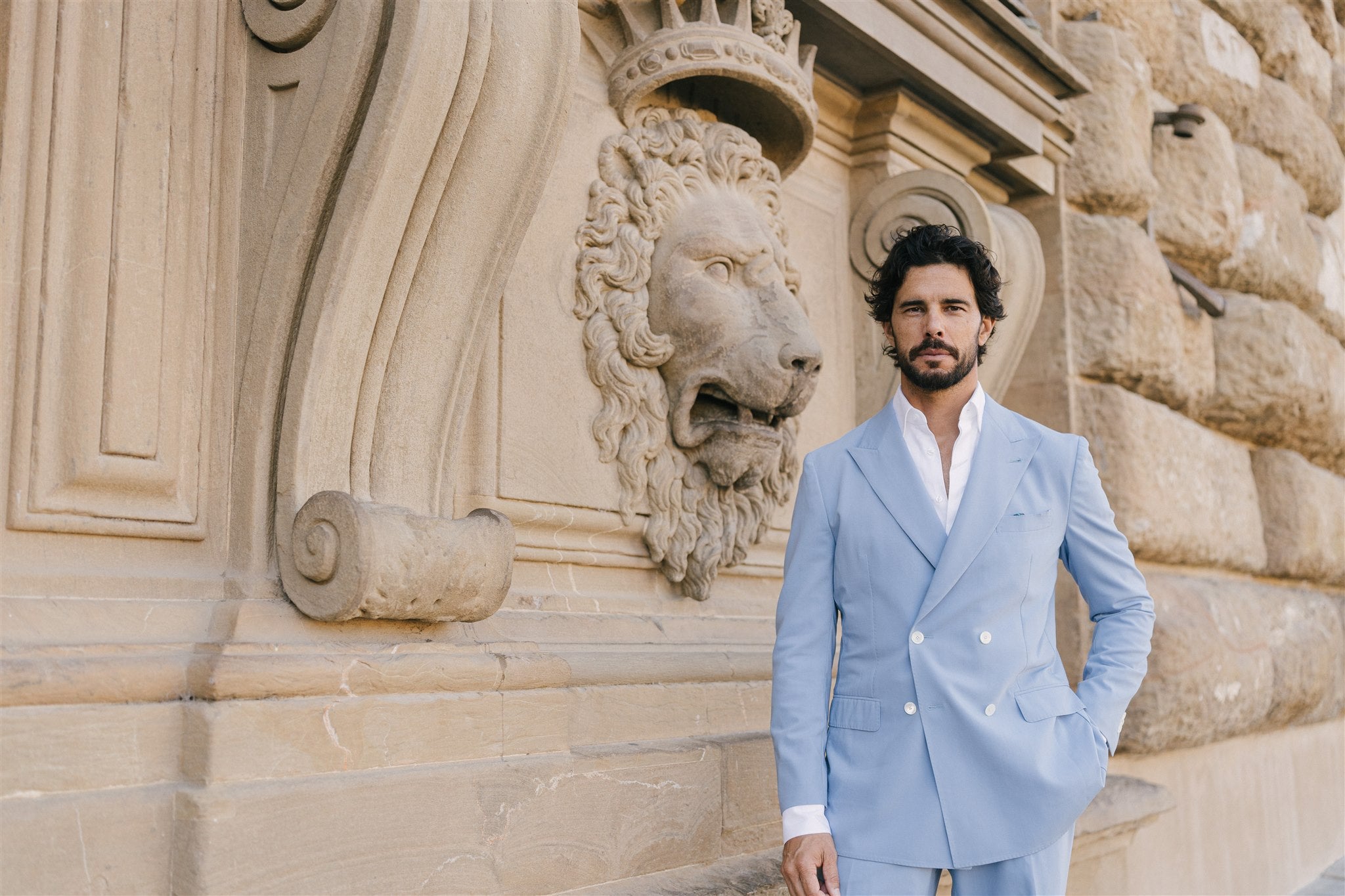 Man in a light blue suit standing in front of a stone wall with lion sculptures