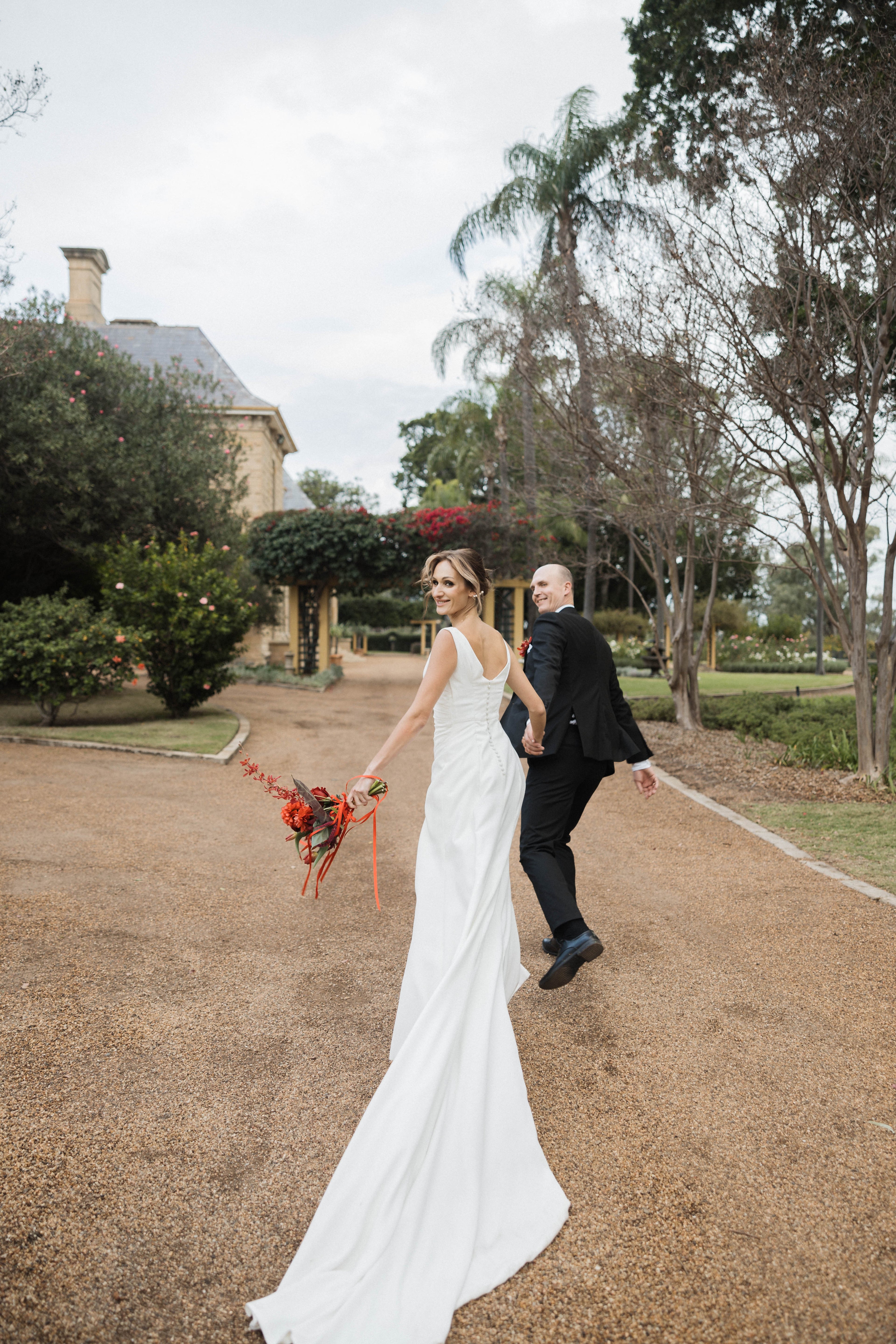 Bride in a white dress holding a bouquet, walking with a man in a black suit on a path in a garden setting.