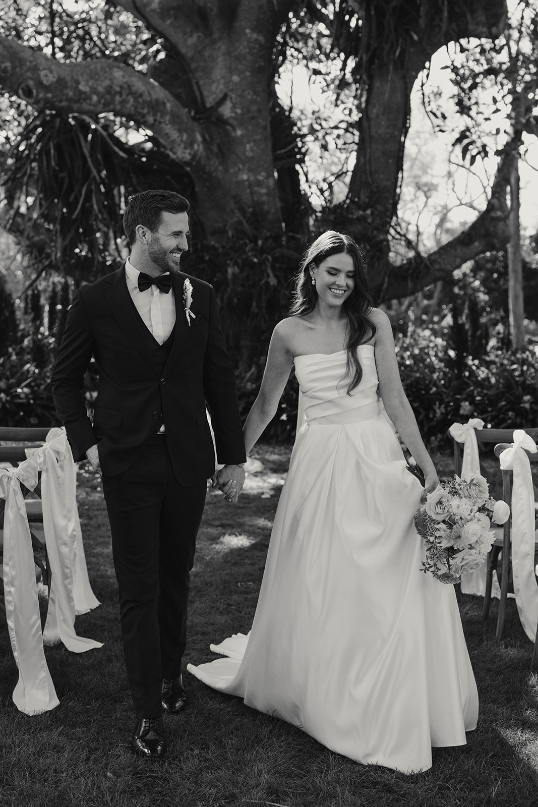 Black and white photo of a couple walking outdoors with floral decorations.