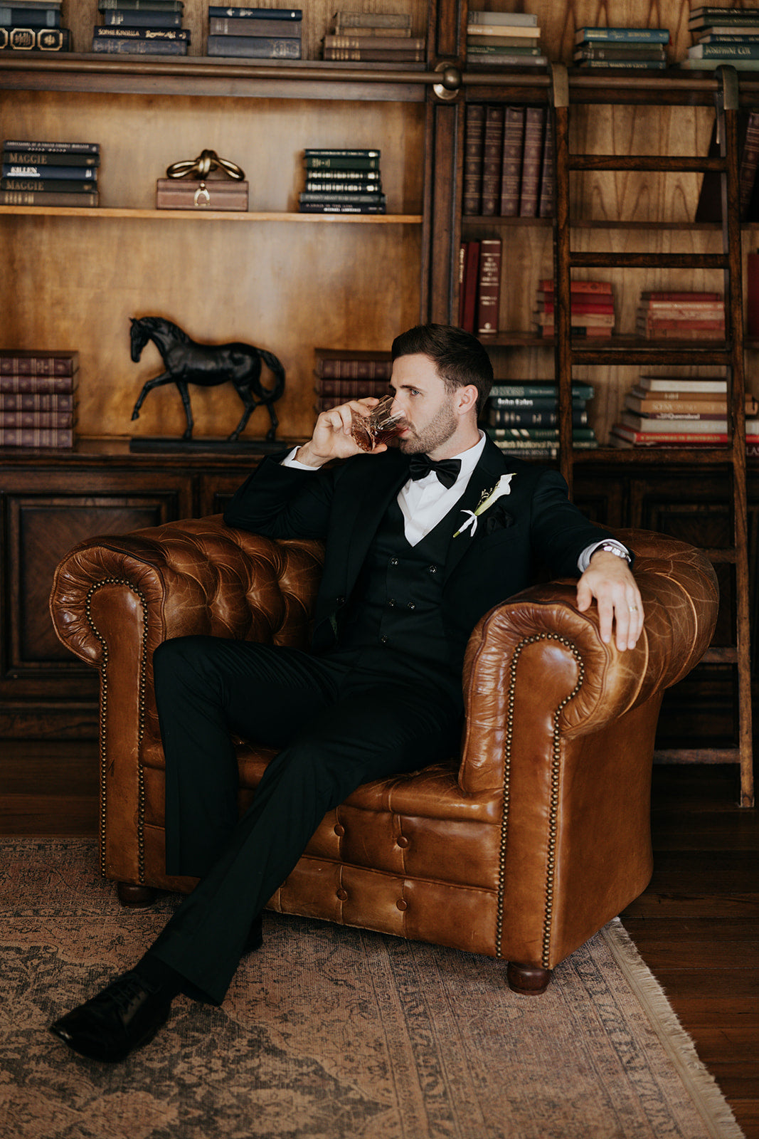 Man in a suit sitting on a brown leather armchair in a library setting.