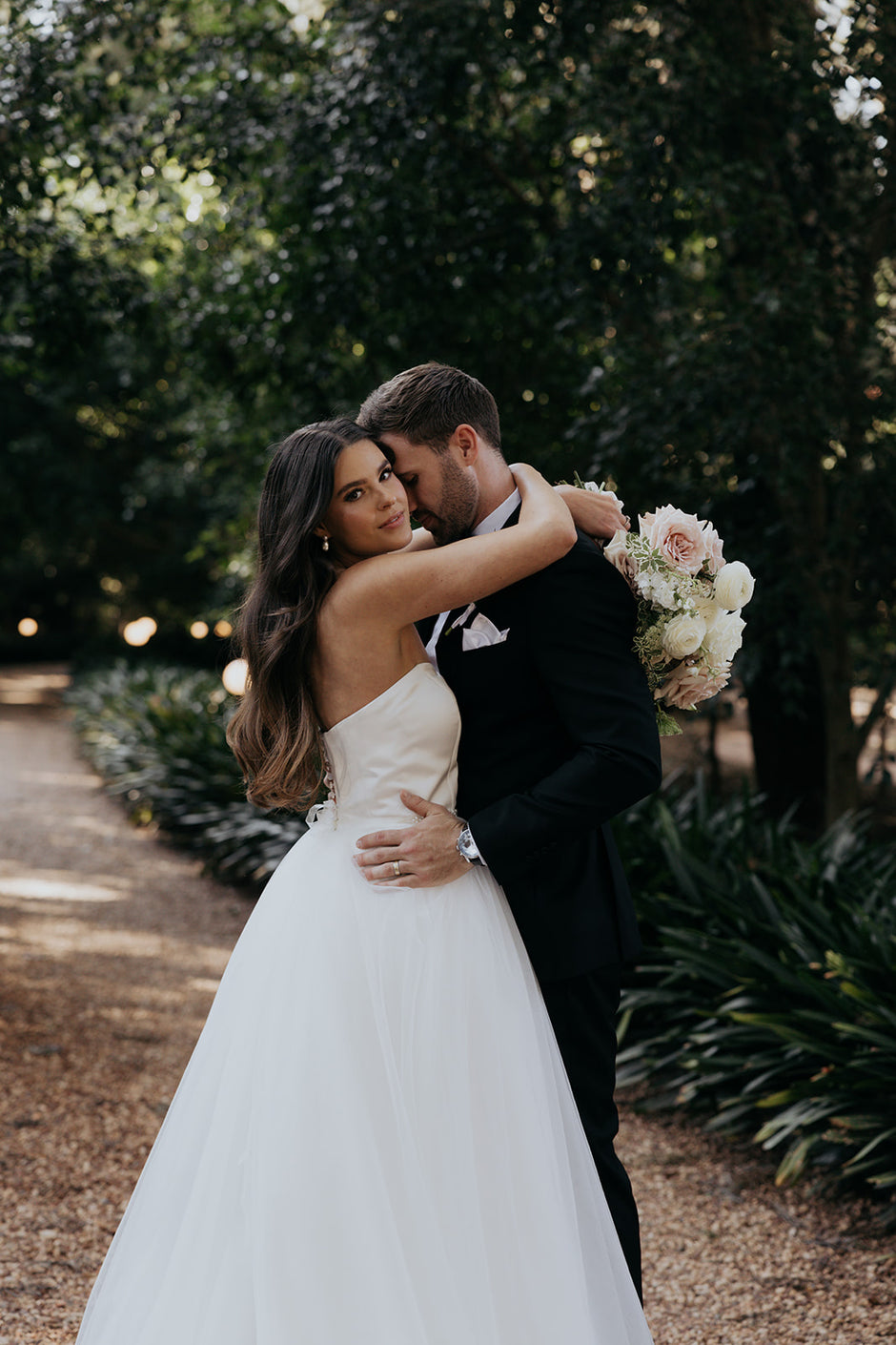 Couple in wedding attire embracing outdoors with a bouquet of flowers.