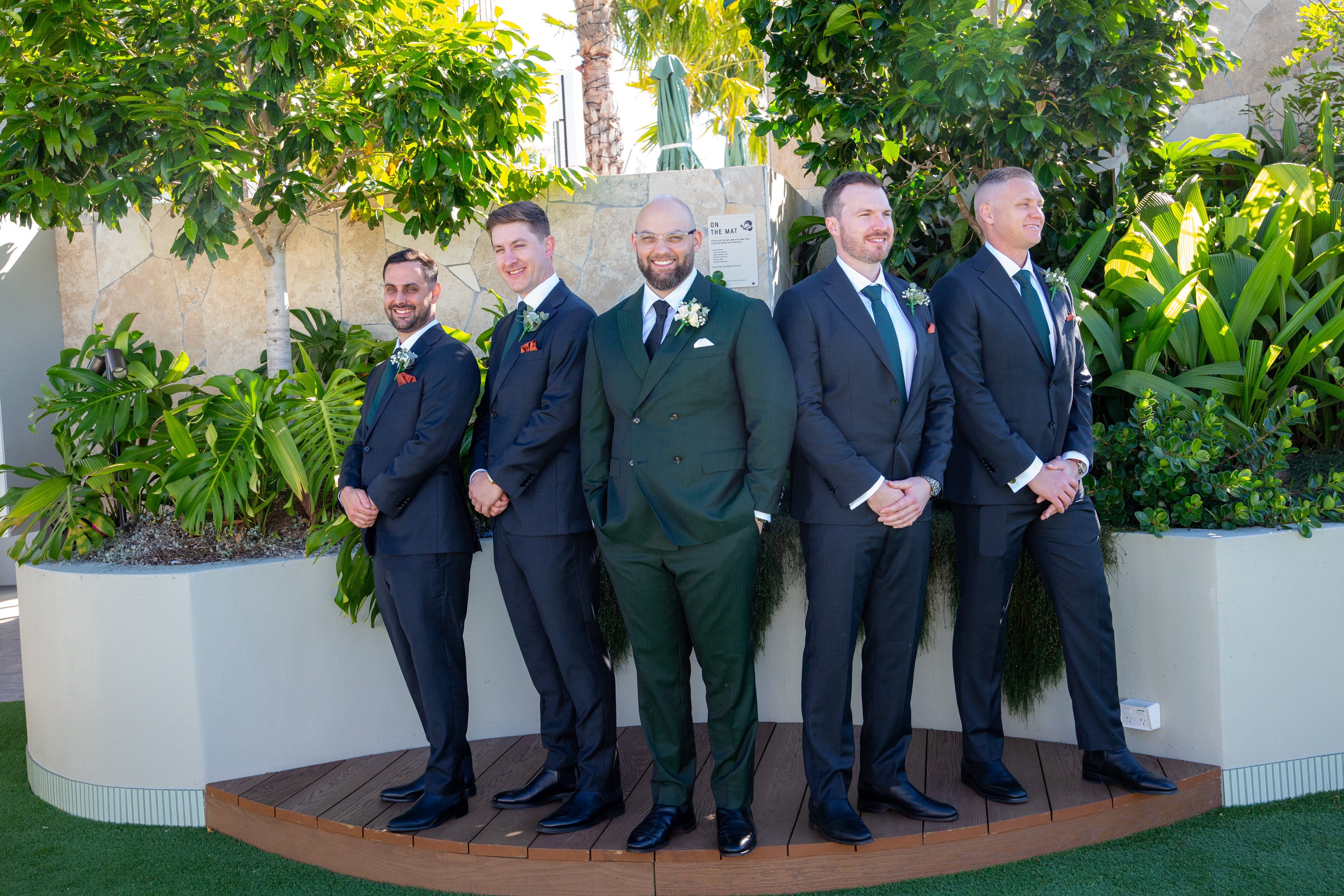 Five men in suits standing outdoors with greenery in the background