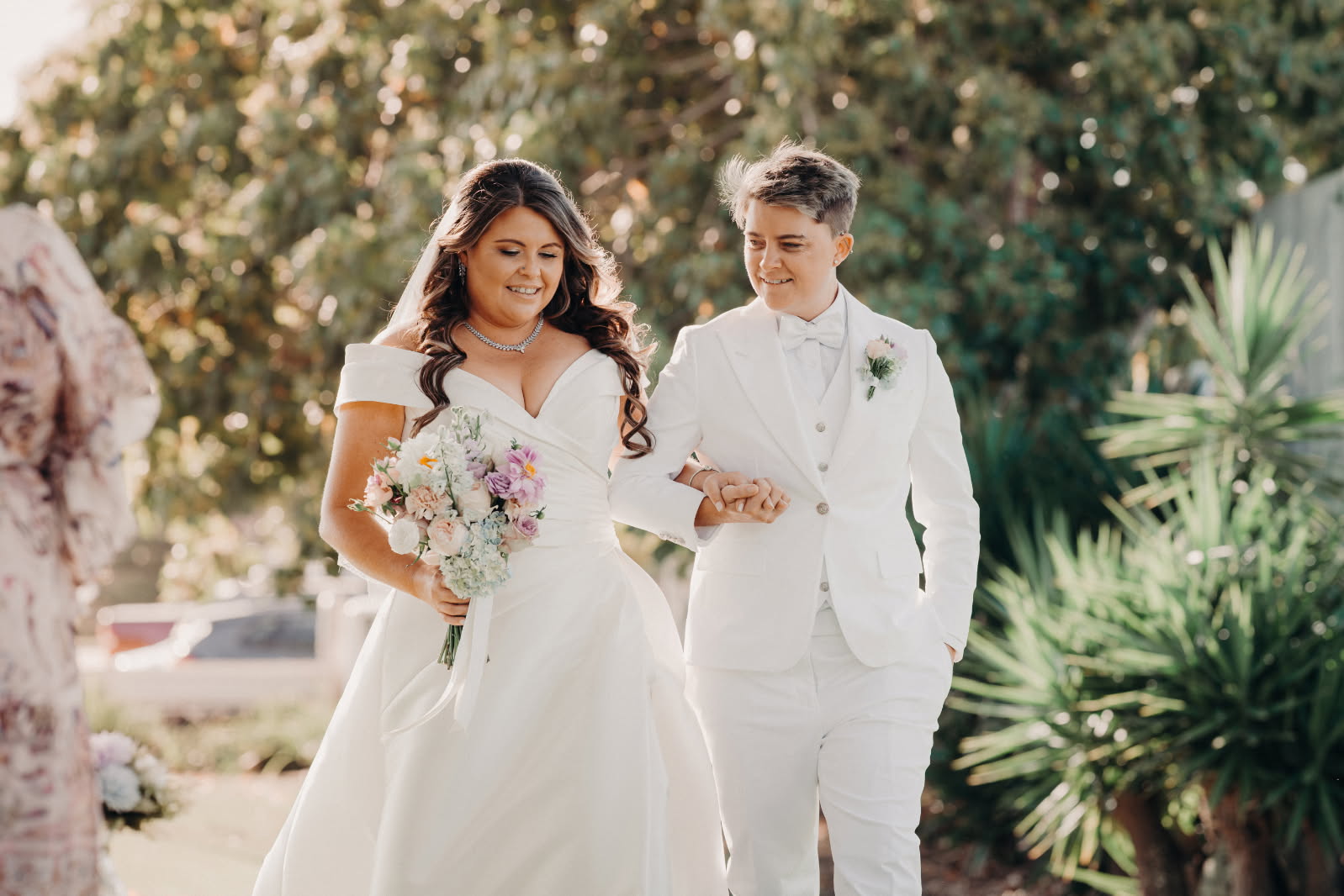 Two people in wedding attire walking together outdoors with greenery in the background