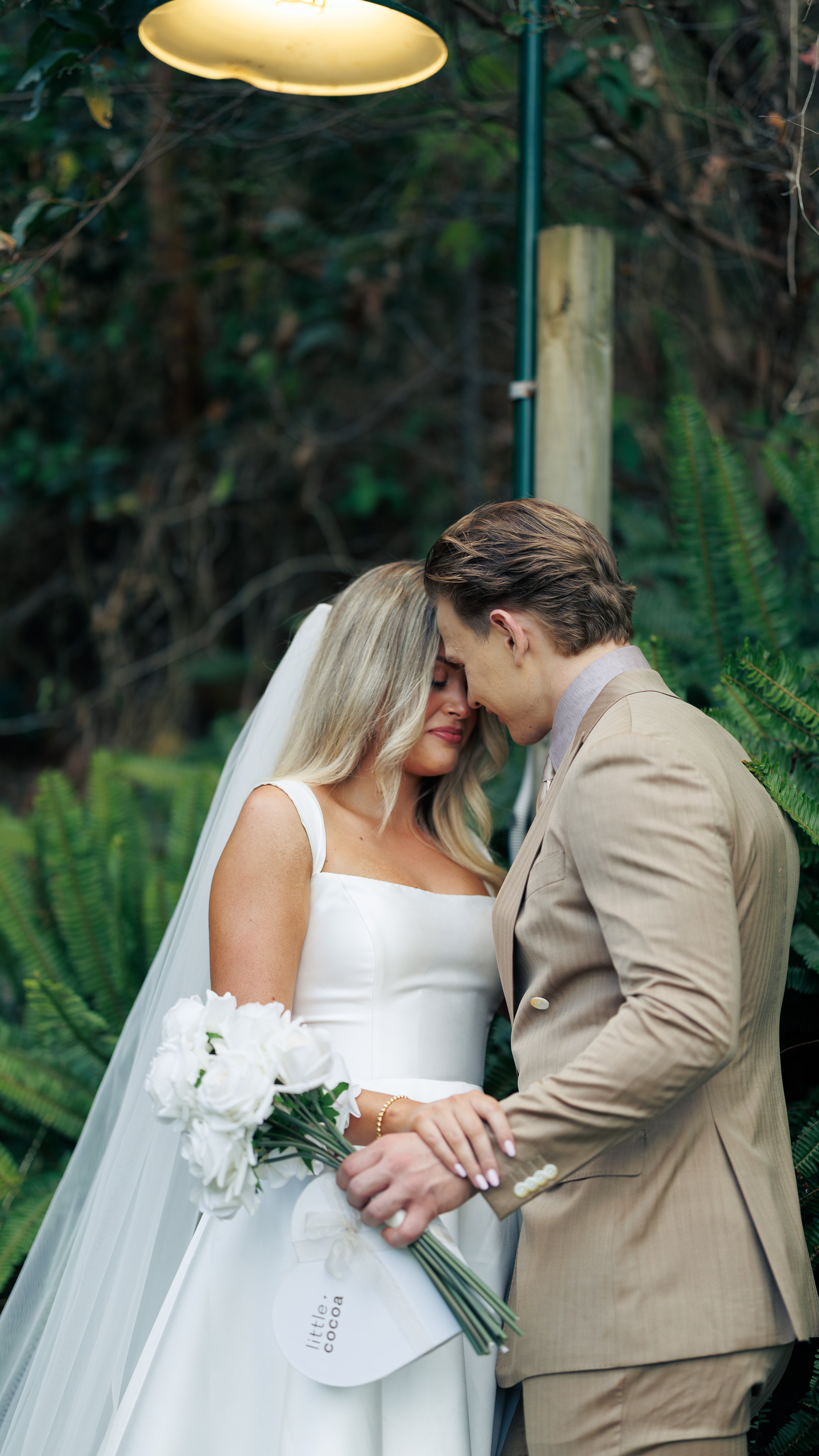Couple in wedding attire standing close together outdoors with greenery in the background