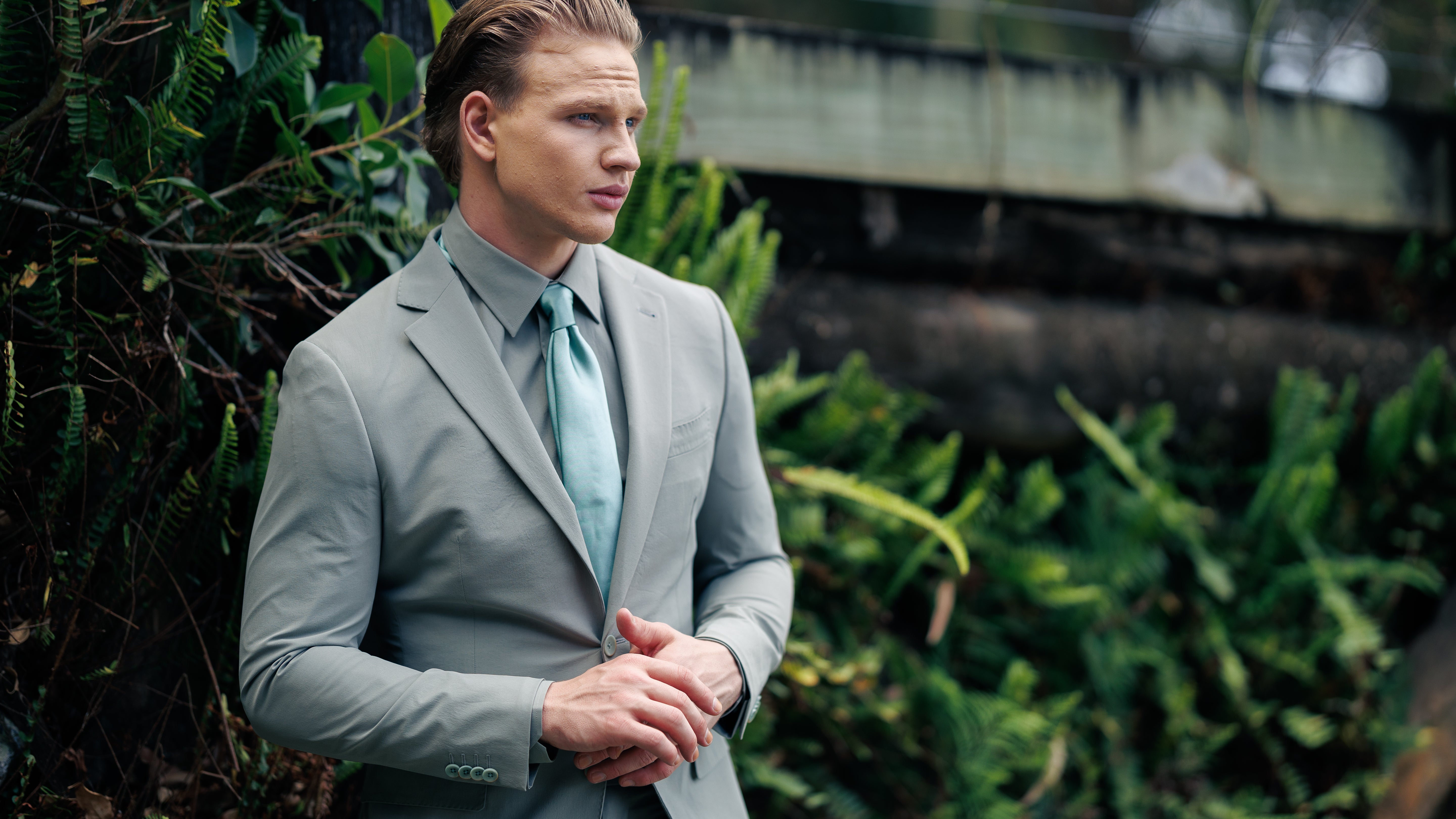 Man in a gray suit standing outdoors with greenery around
