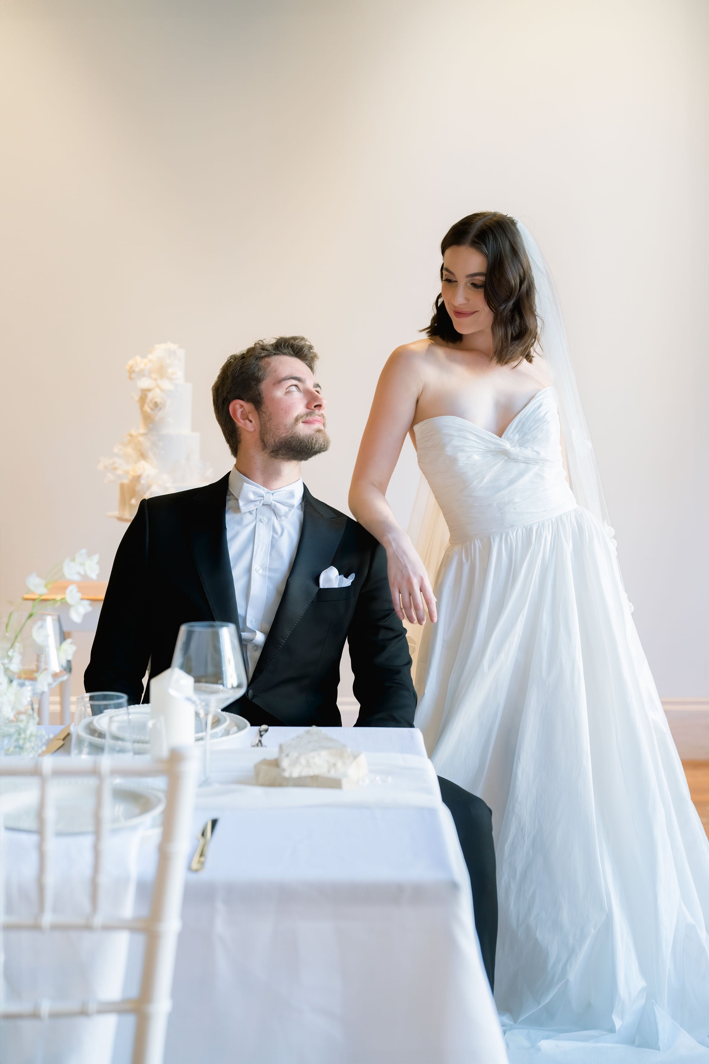 Man and woman in wedding attire standing near a wedding cake in a celebratory setting.