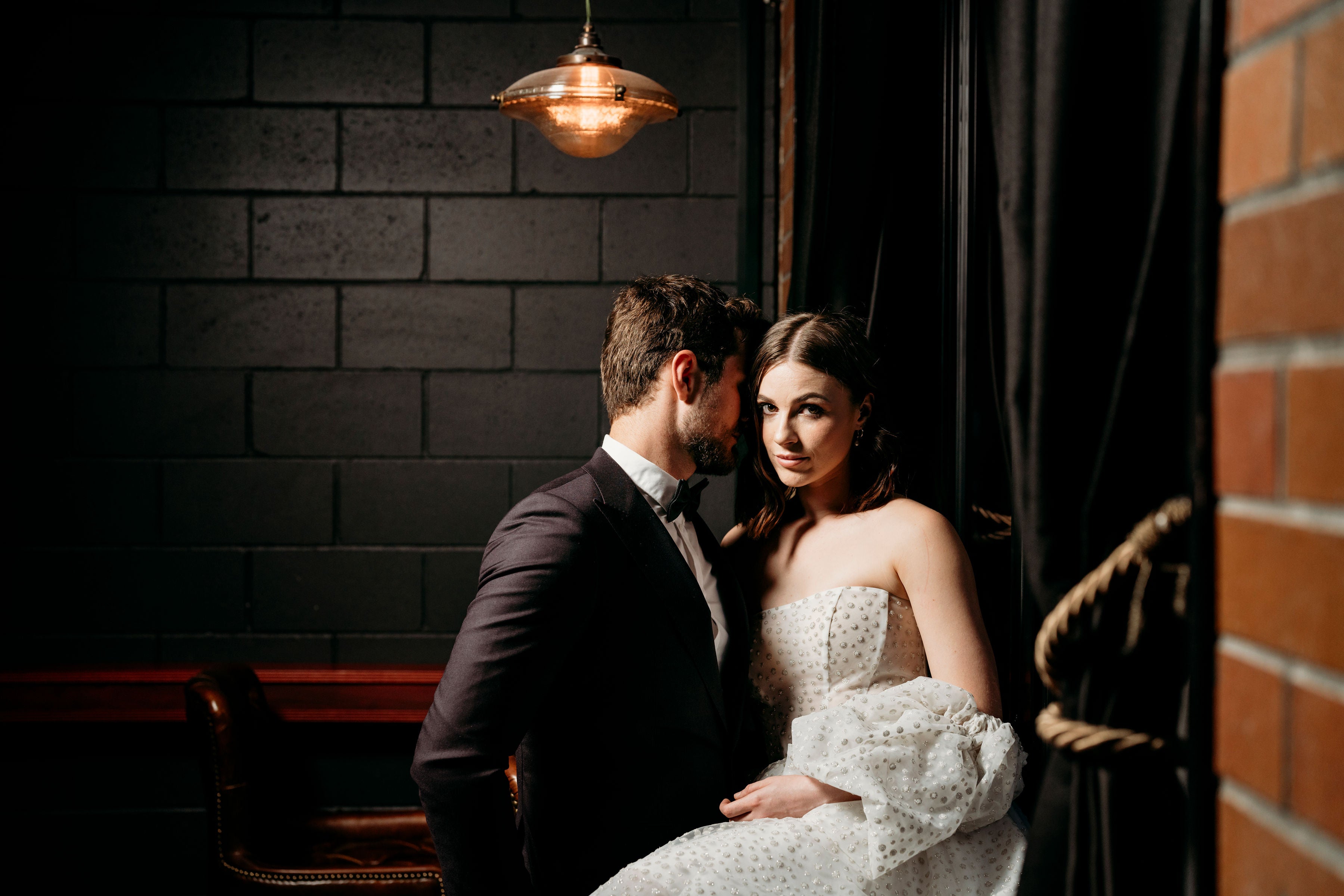 Couple in formal attire sitting close together against a brick wall.