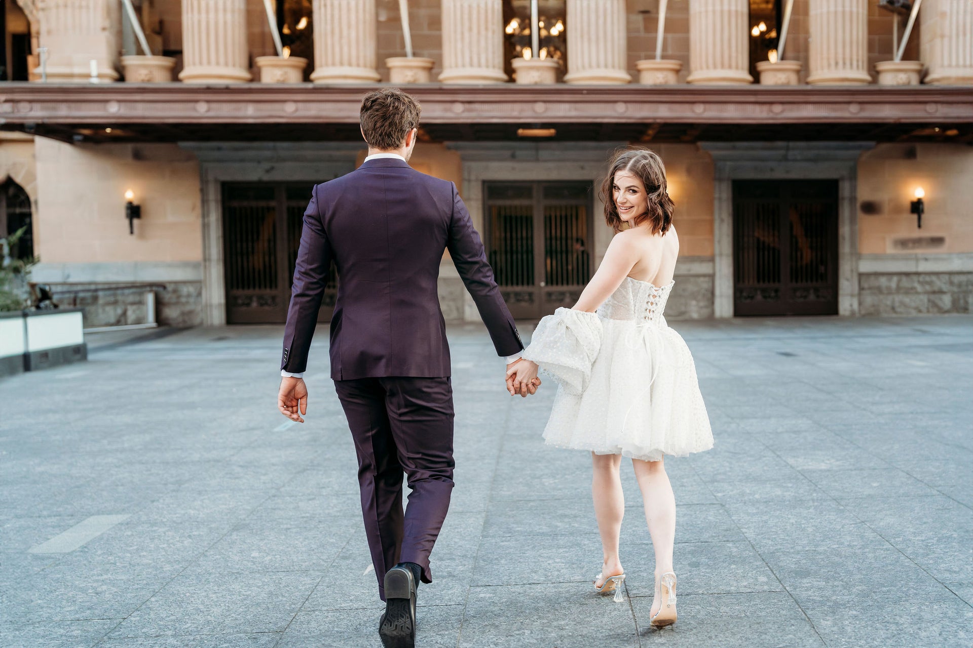Man and woman holding hands in formal attire in front of a classical building.