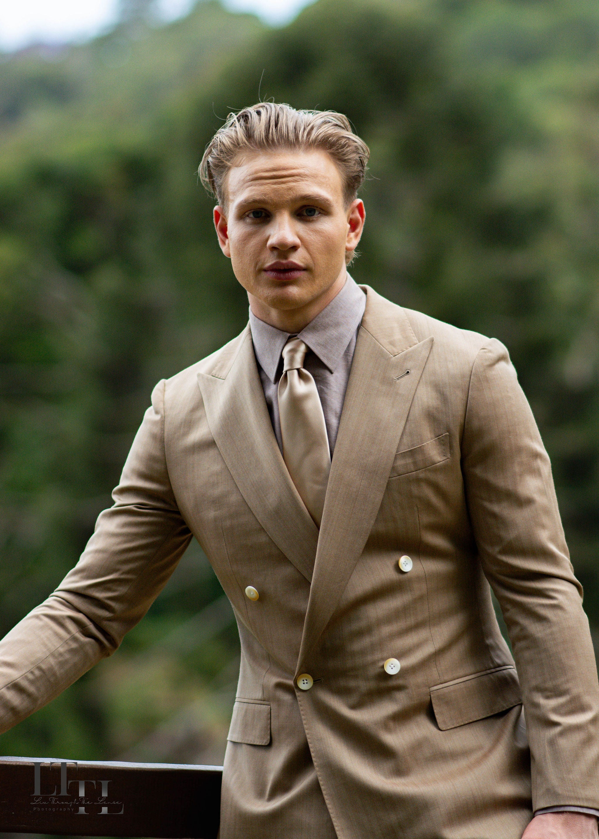 Man in a beige suit standing outdoors with greenery in the background