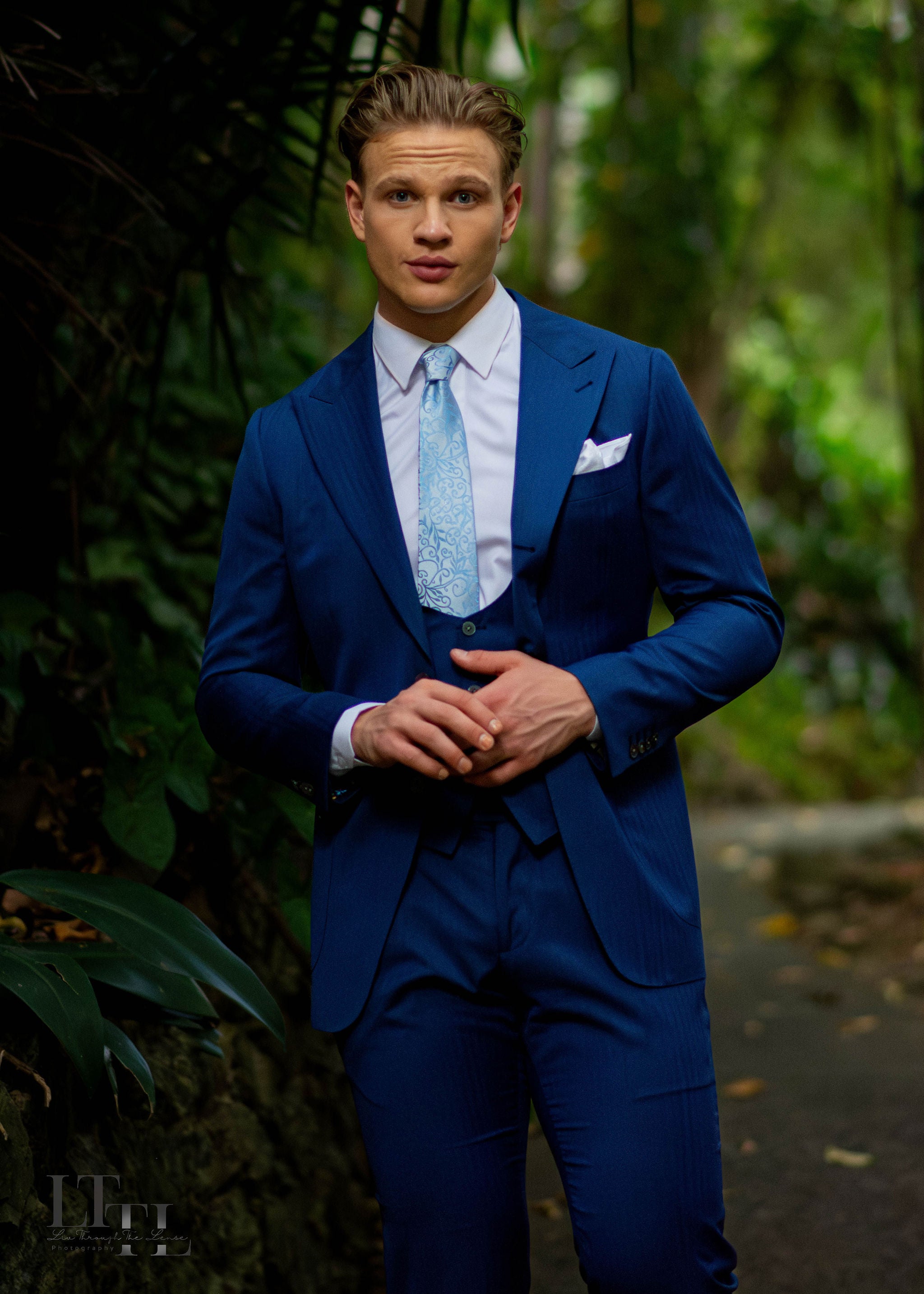 Man in a blue suit standing outdoors with greenery in the background