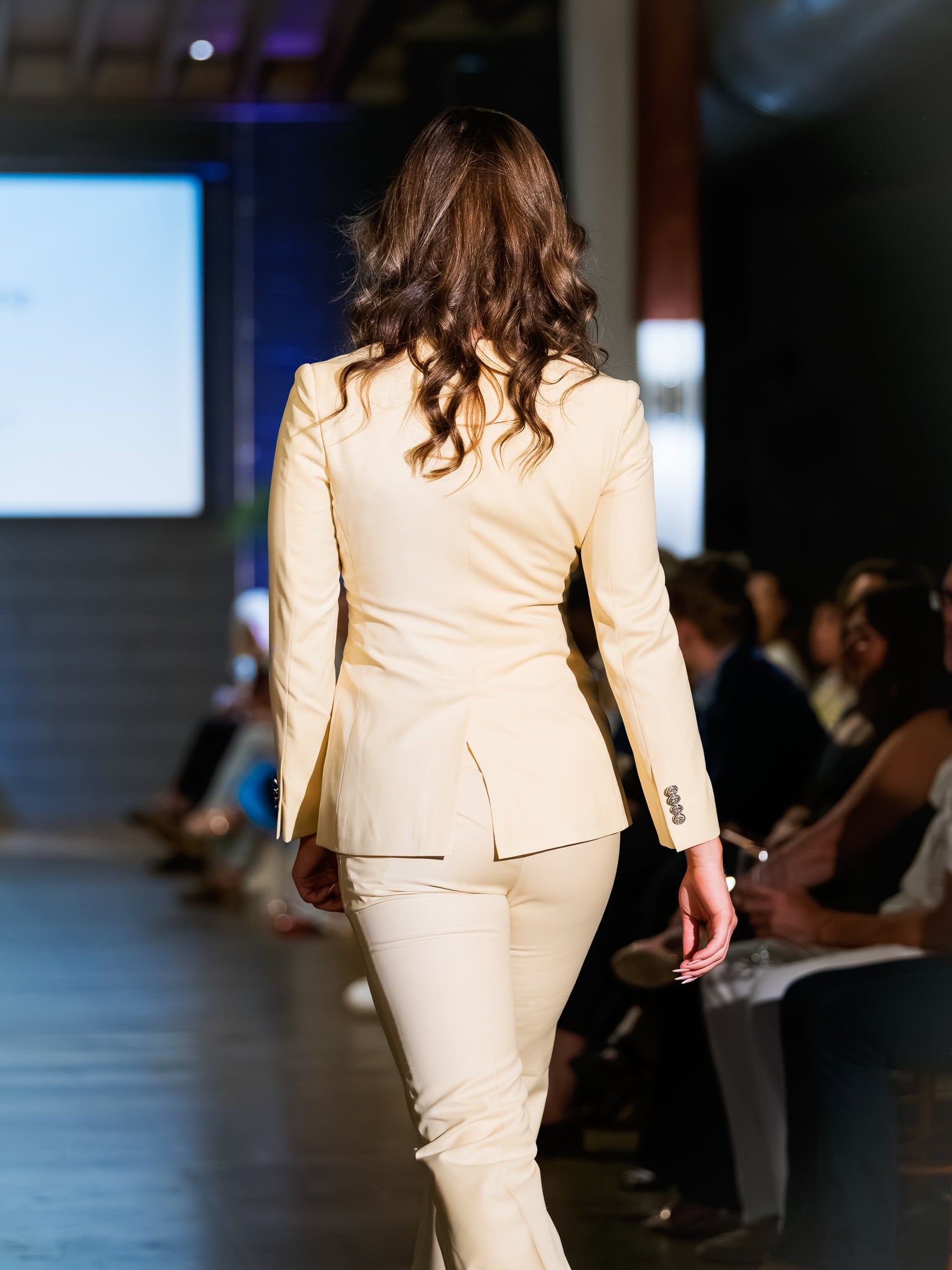 Woman in a beige suit walking on a runway with an audience in the background.