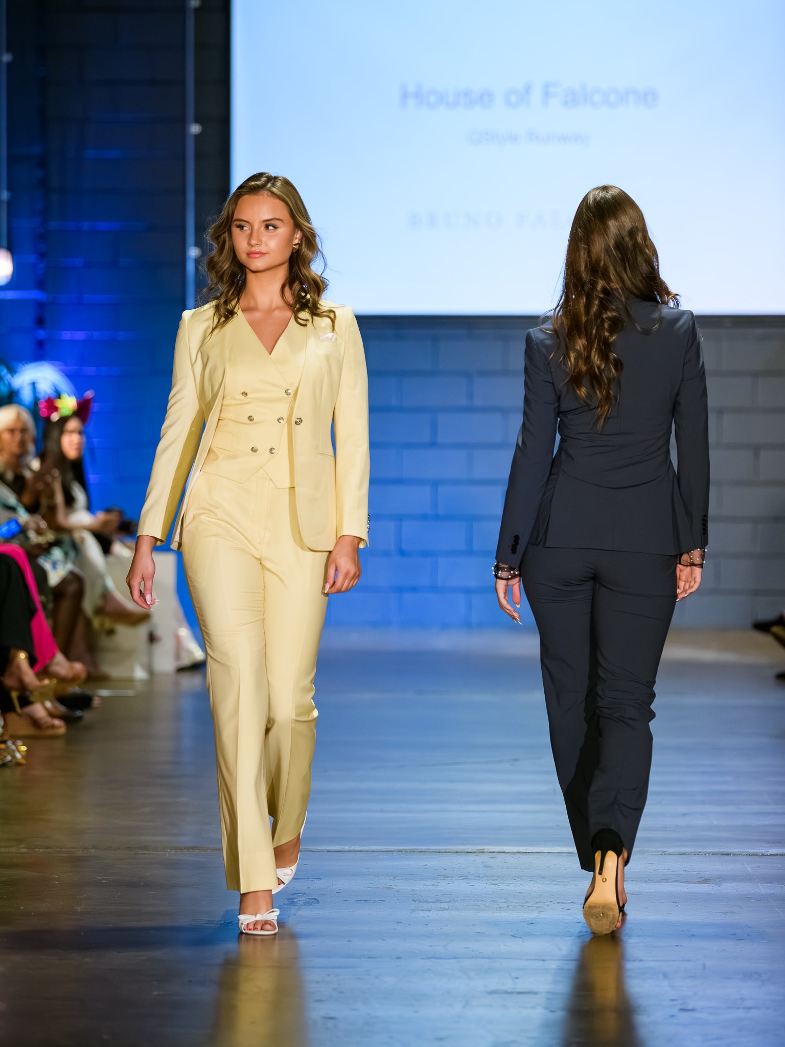 Two models walking down a runway wearing suits, with a screen displaying 'House of Falcone' in the background.