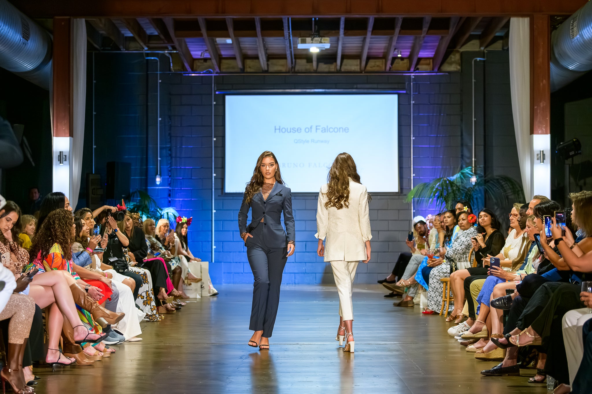 Two models walking down a runway at a fashion show with an audience on either side.