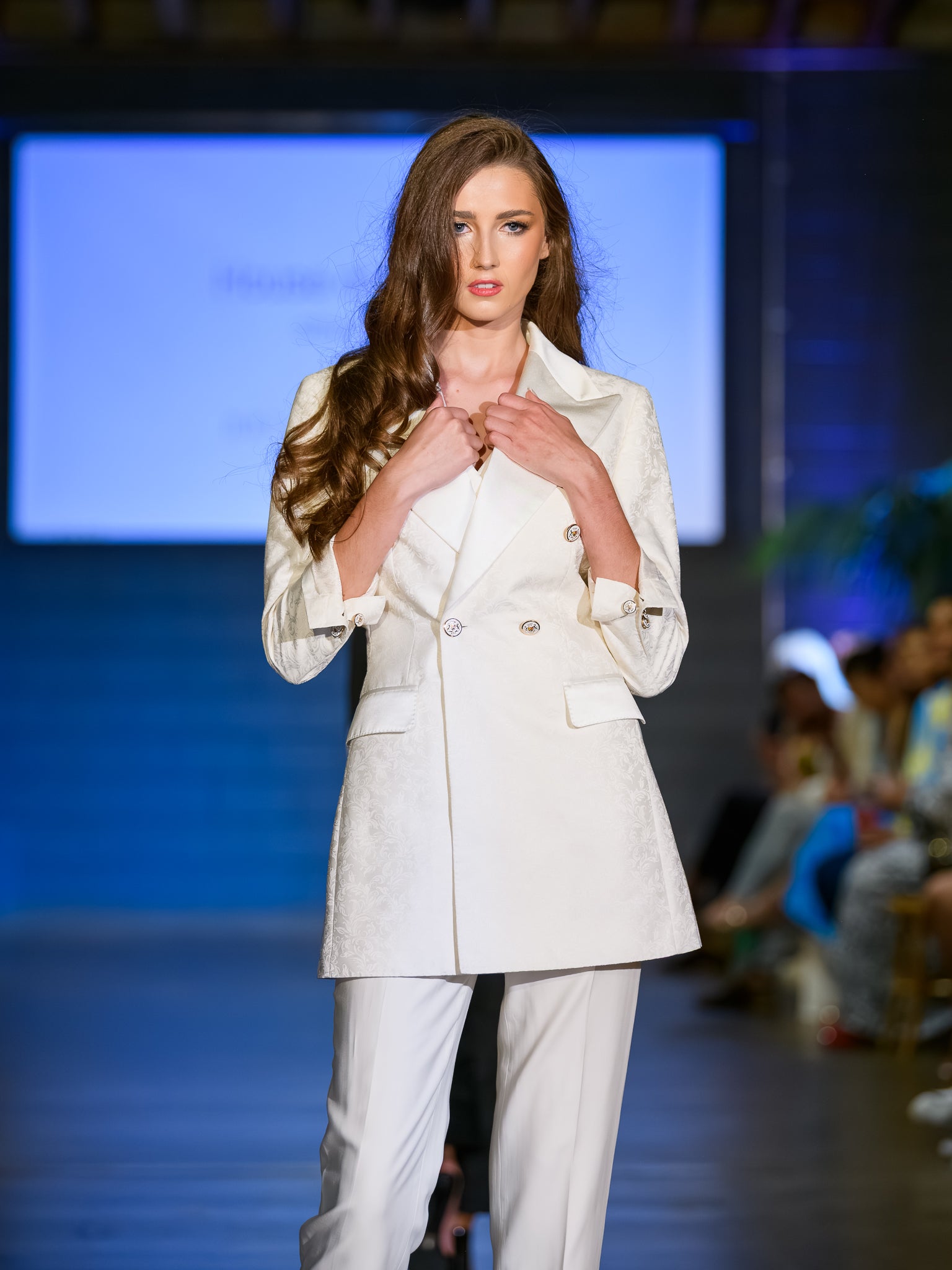 Model wearing a white suit on a runway with a blue screen background