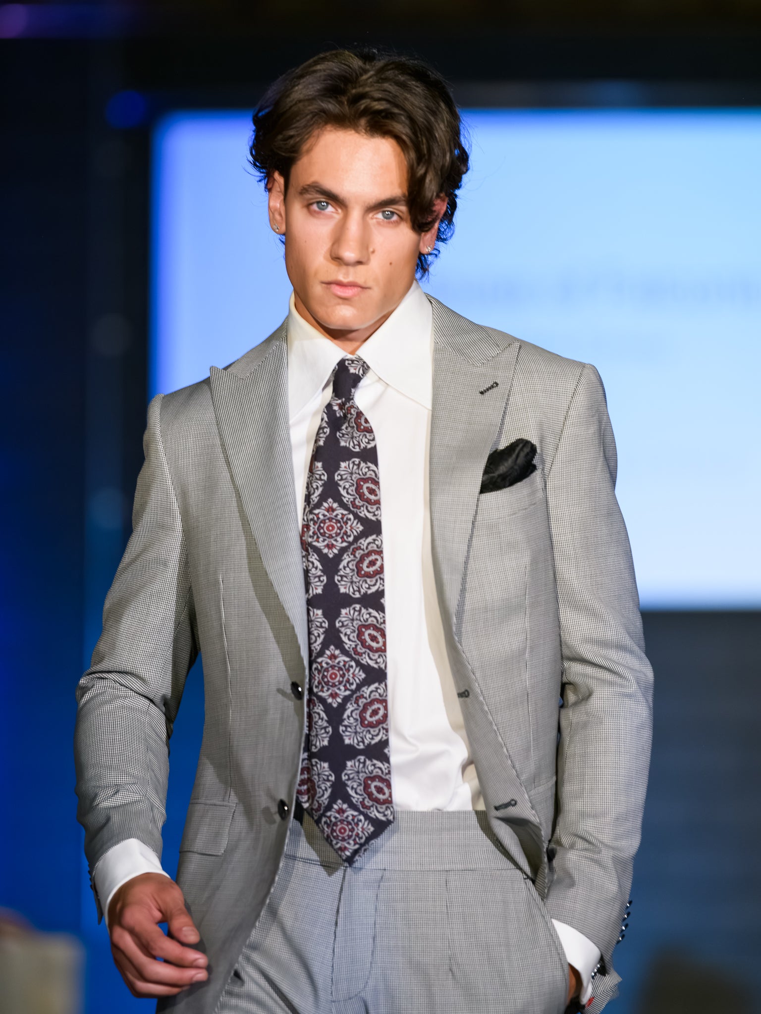 Man in a gray suit with a patterned tie walking on a runway.