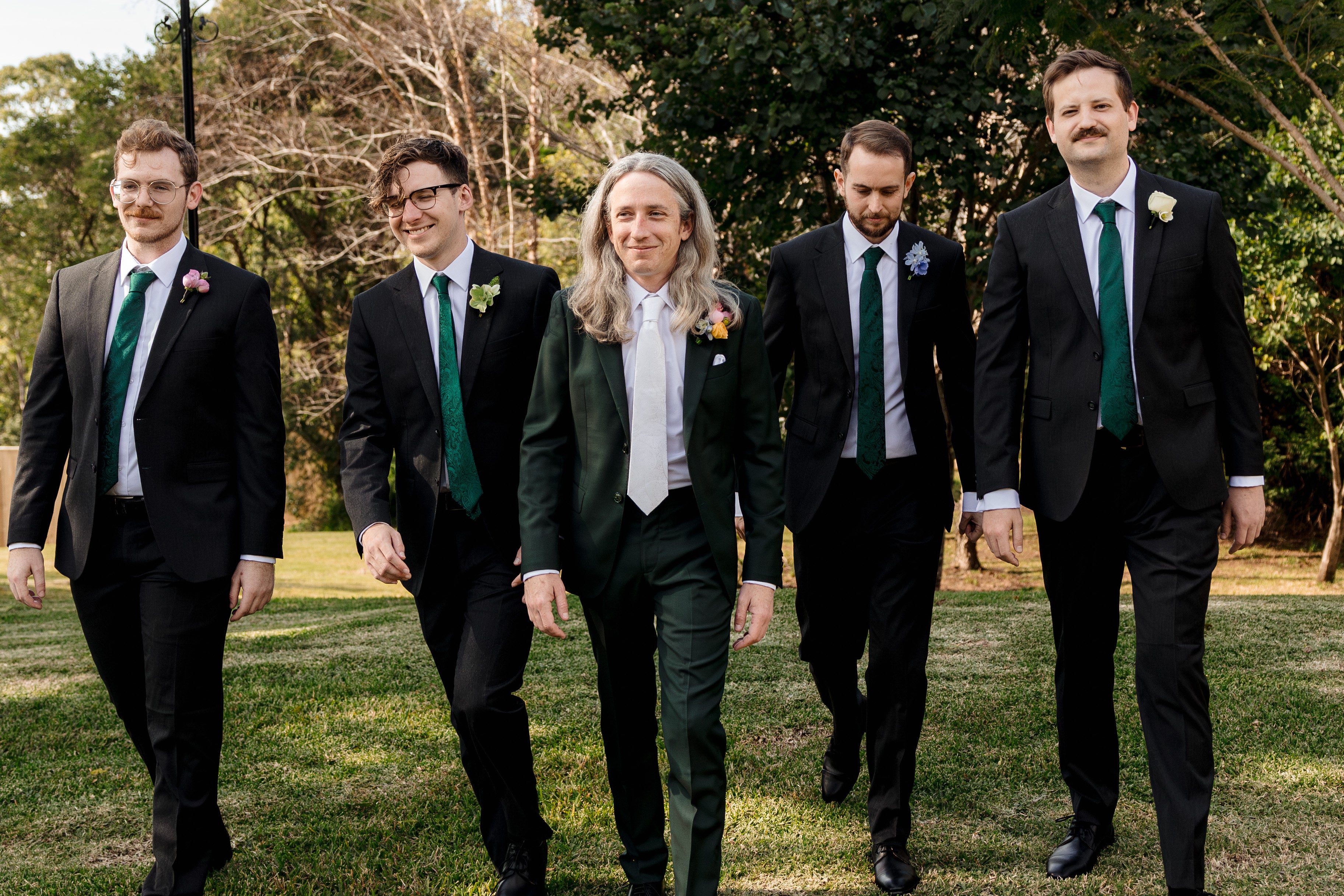 Five men in suits walking outdoors with trees in the background