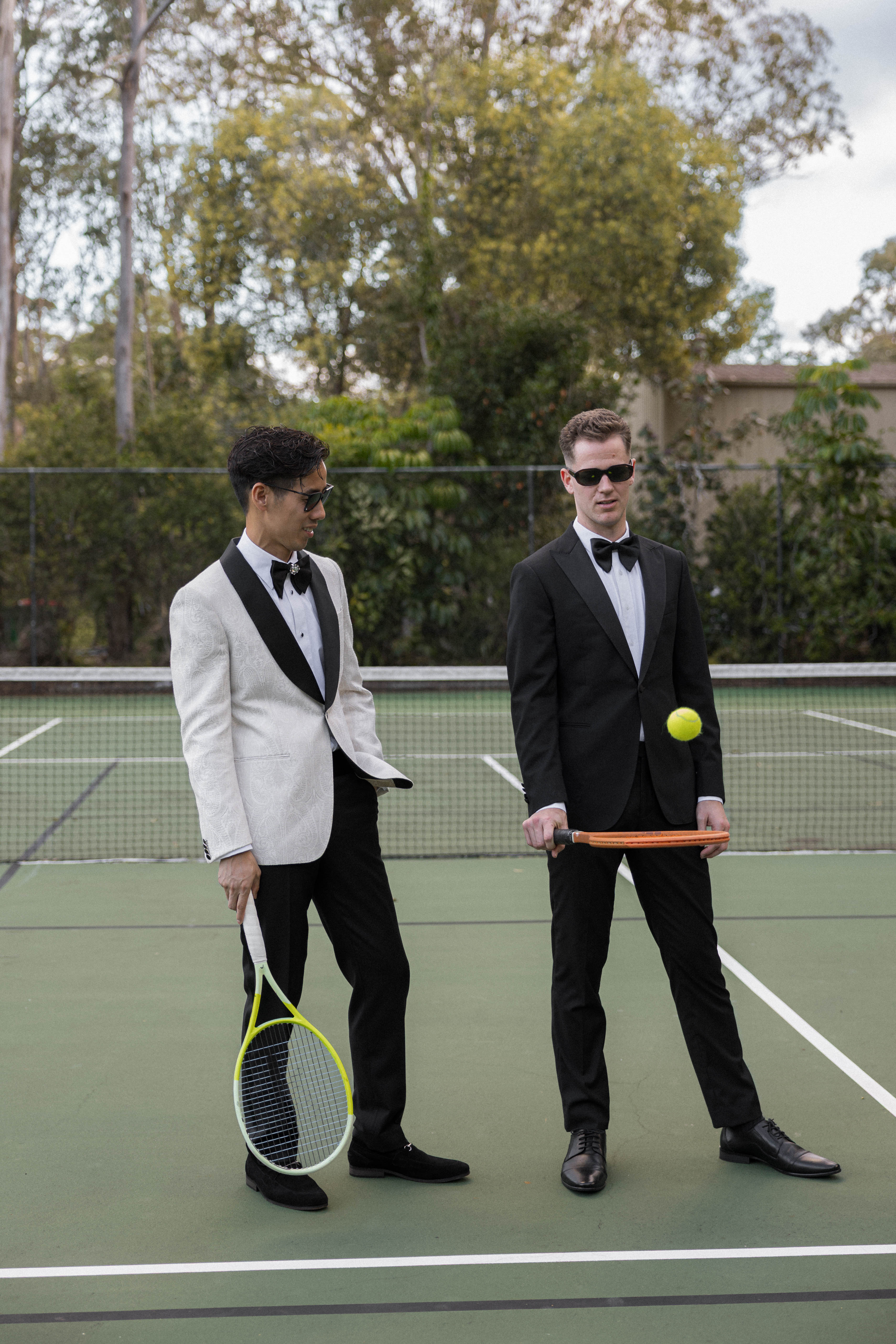 Two men in formal wear on a tennis court holding tennis rackets and a ball.