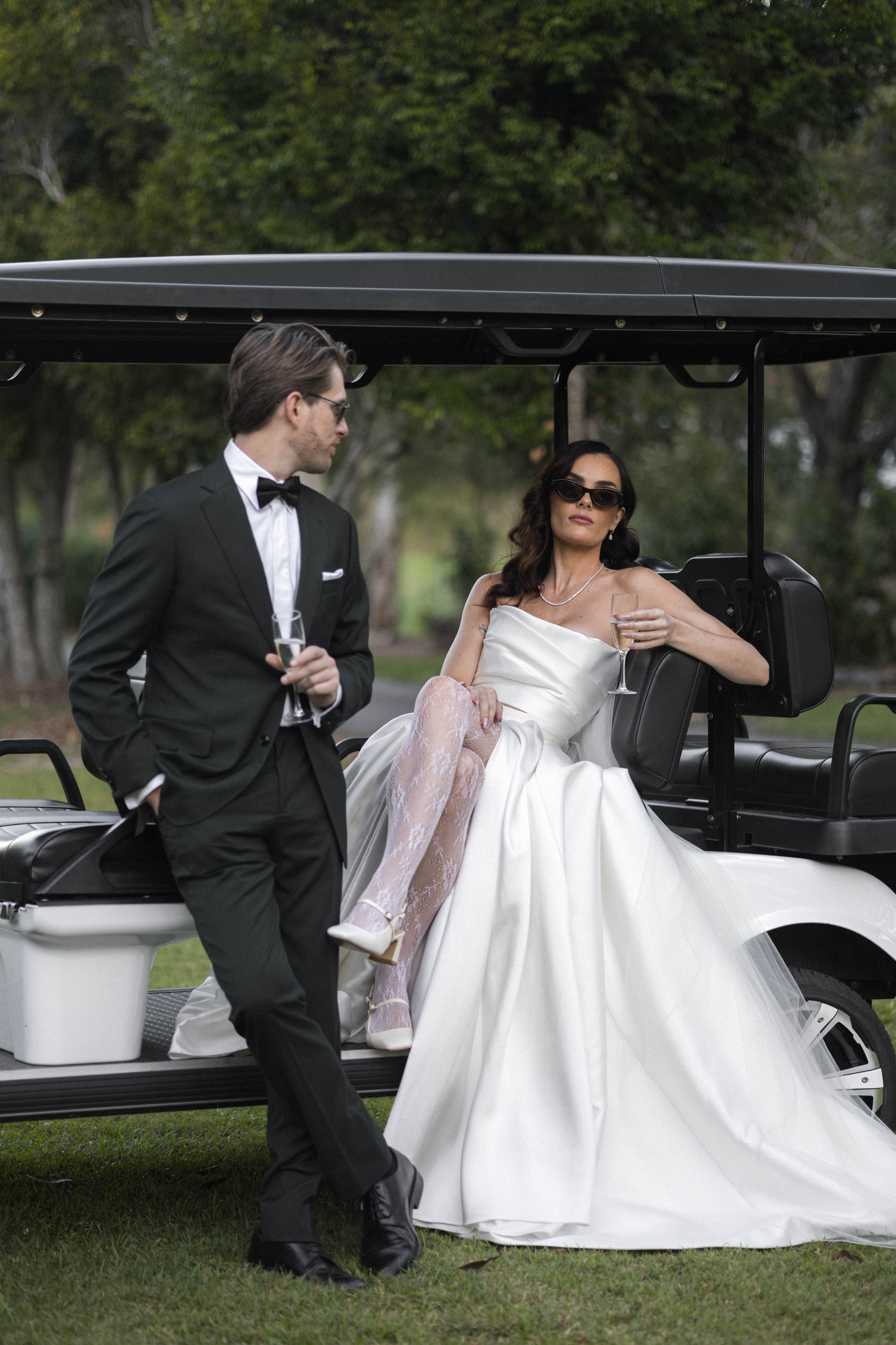 Man in tuxedo and woman in white dress standing next to a golf cart in a park setting.