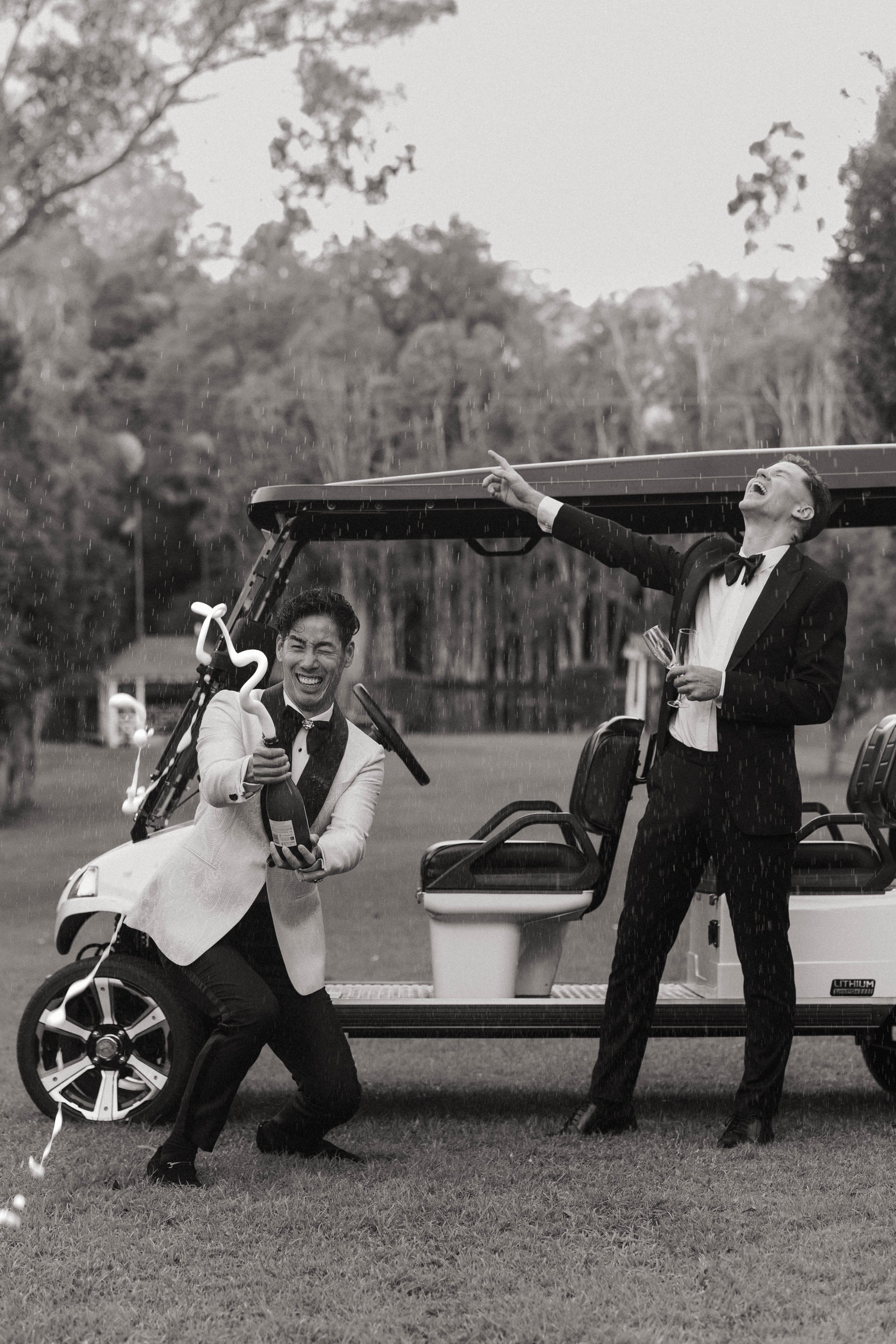 Two men in formal attire standing next to a golf cart on a grassy area with trees in the background.