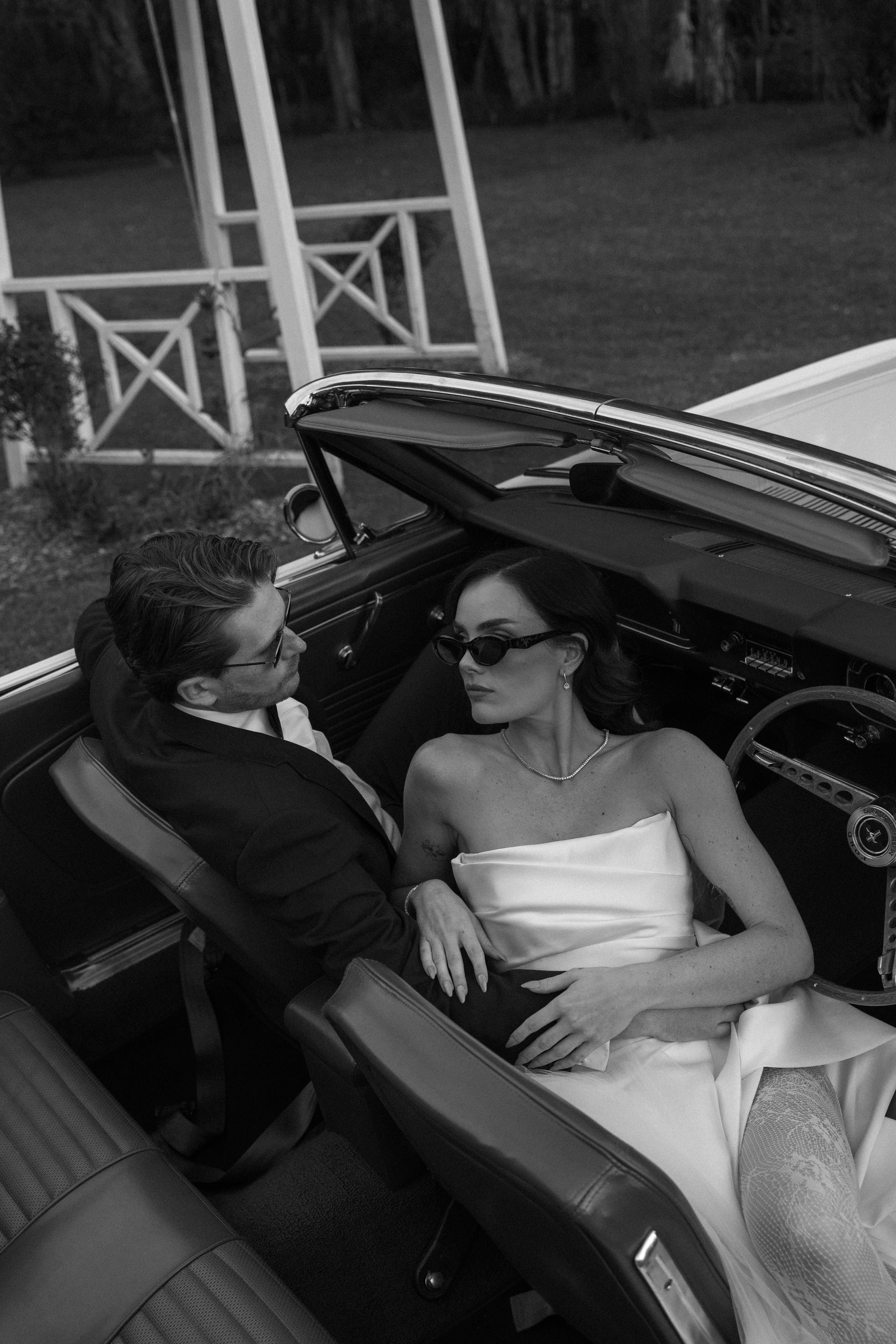 Two people sitting in an open-top car, one in formal attire, in a black and white photo.