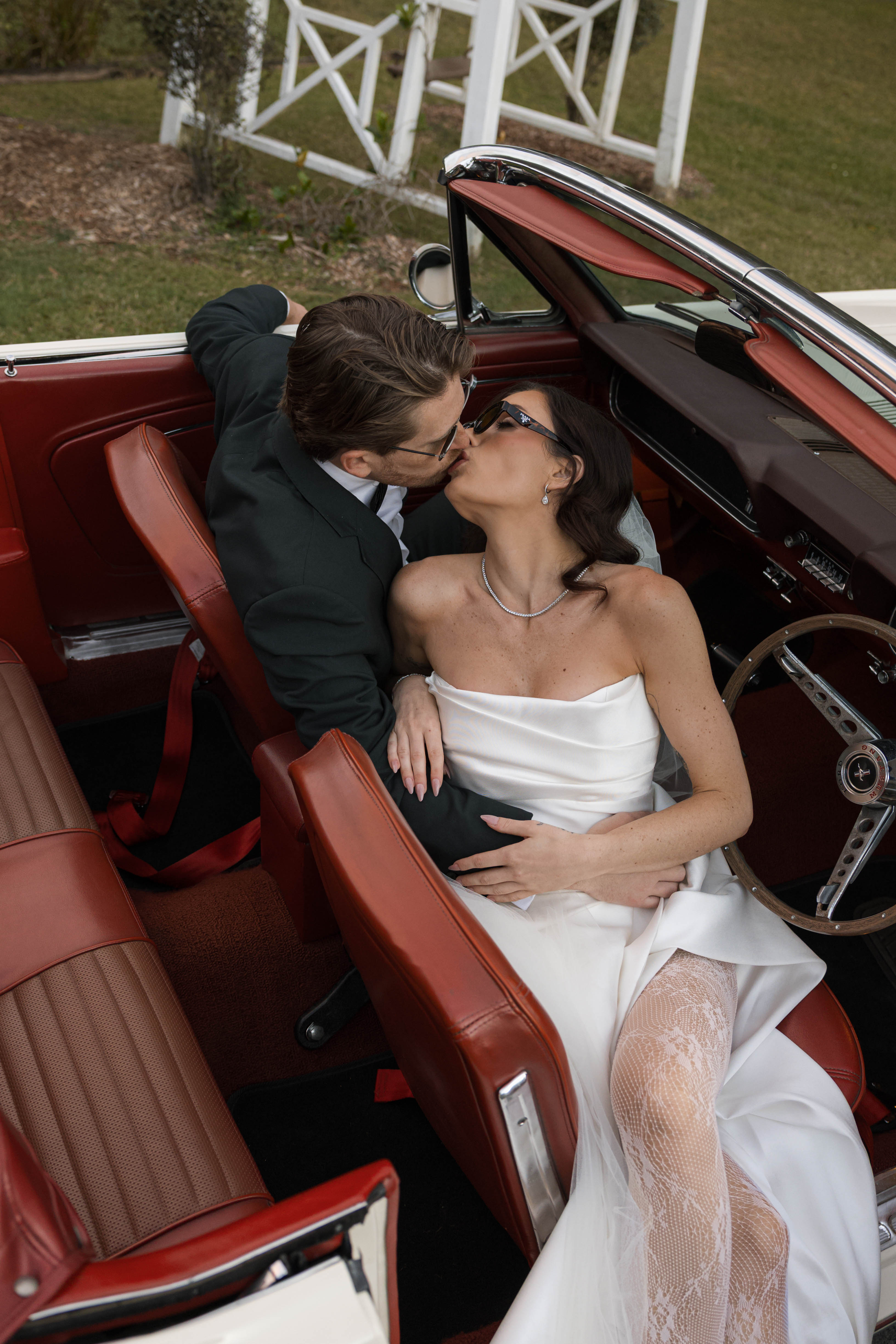 Couple in a red convertible car, bride in white dress and groom in suit, kissing.