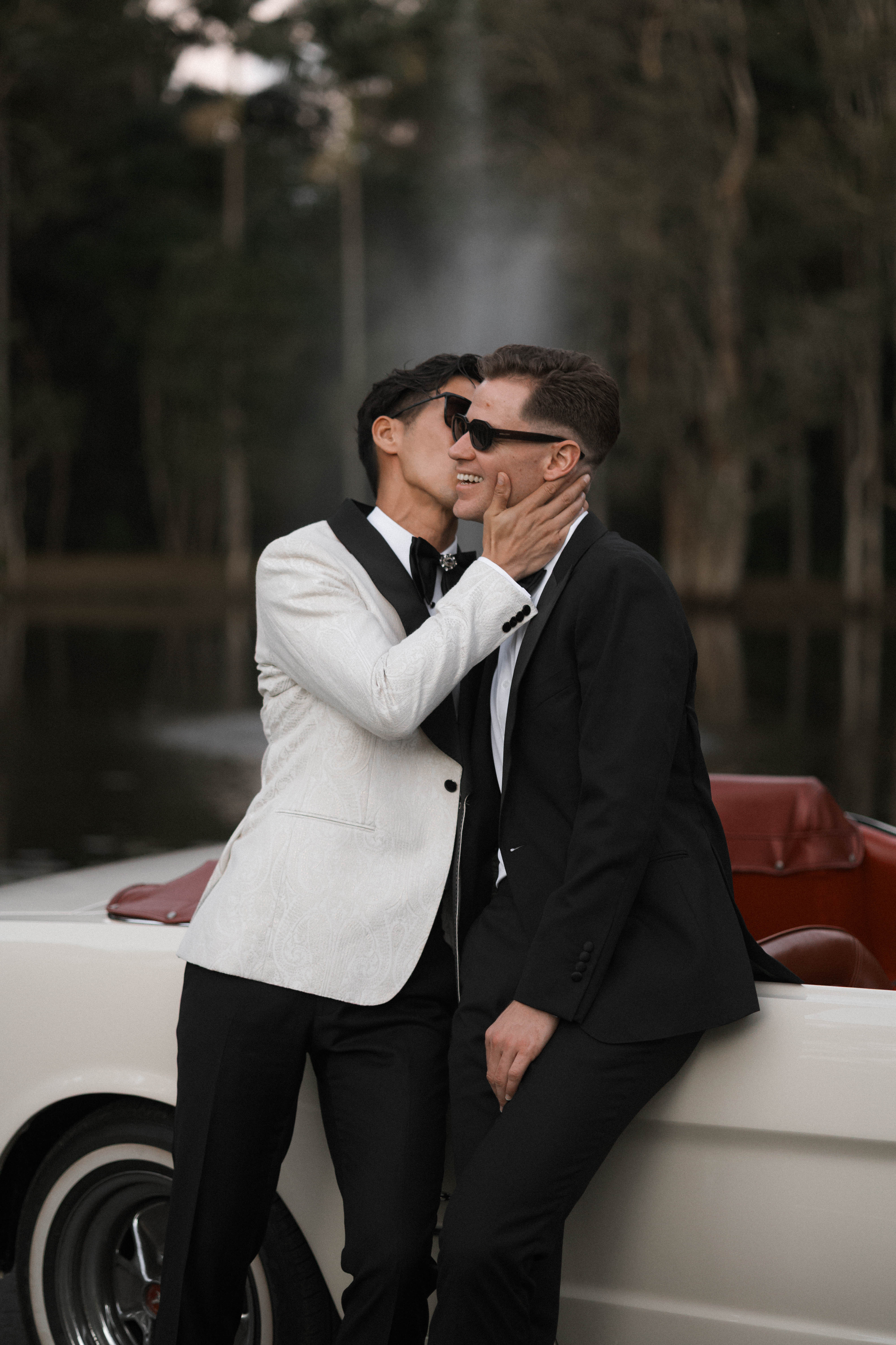 Two men in formal attire embracing on a car with a blurred natural background