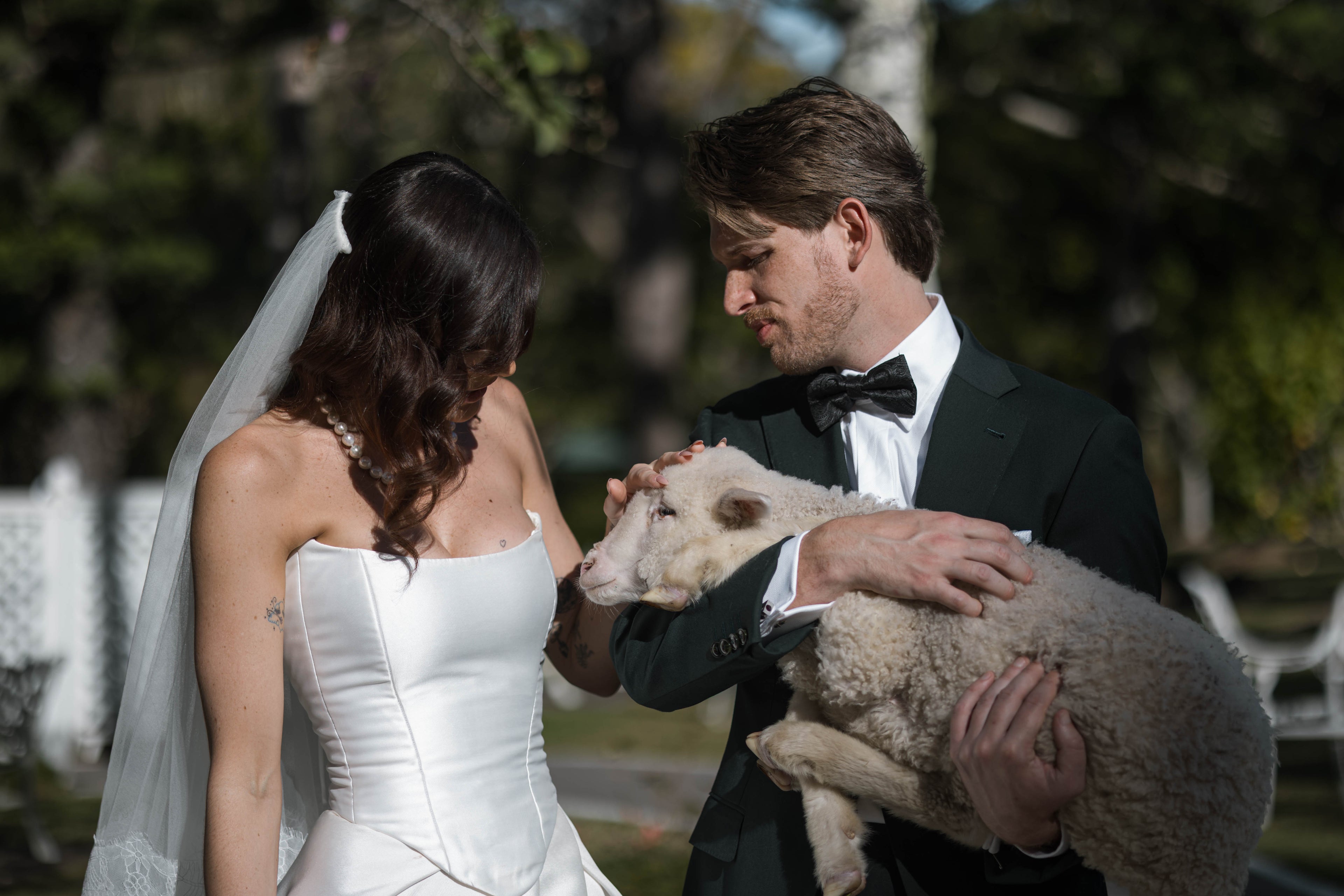 Man in tuxedo holding a sheep next to a woman in a wedding dress outdoors.