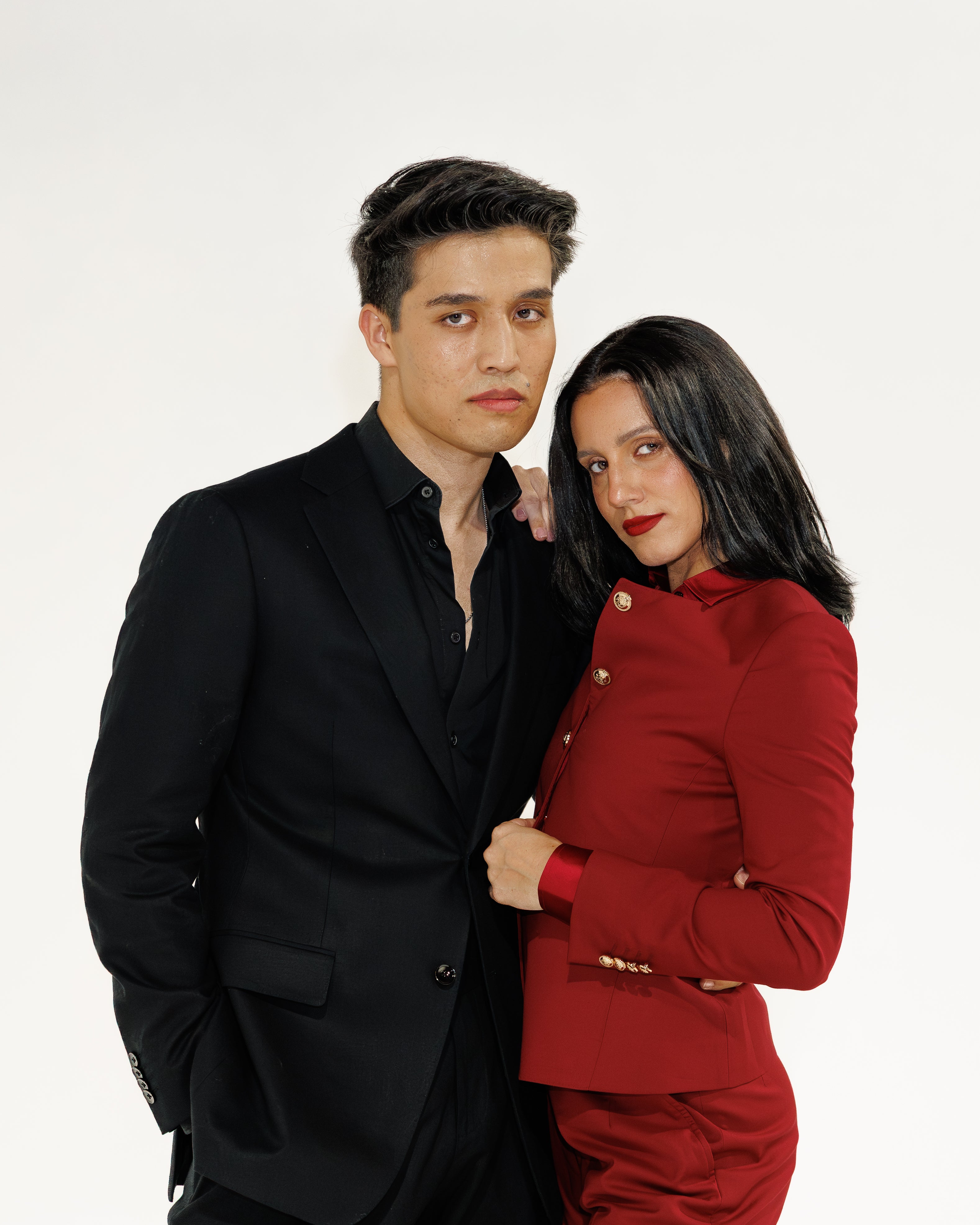 Man in black linen suit and woman in red outfit standing close together on a white background