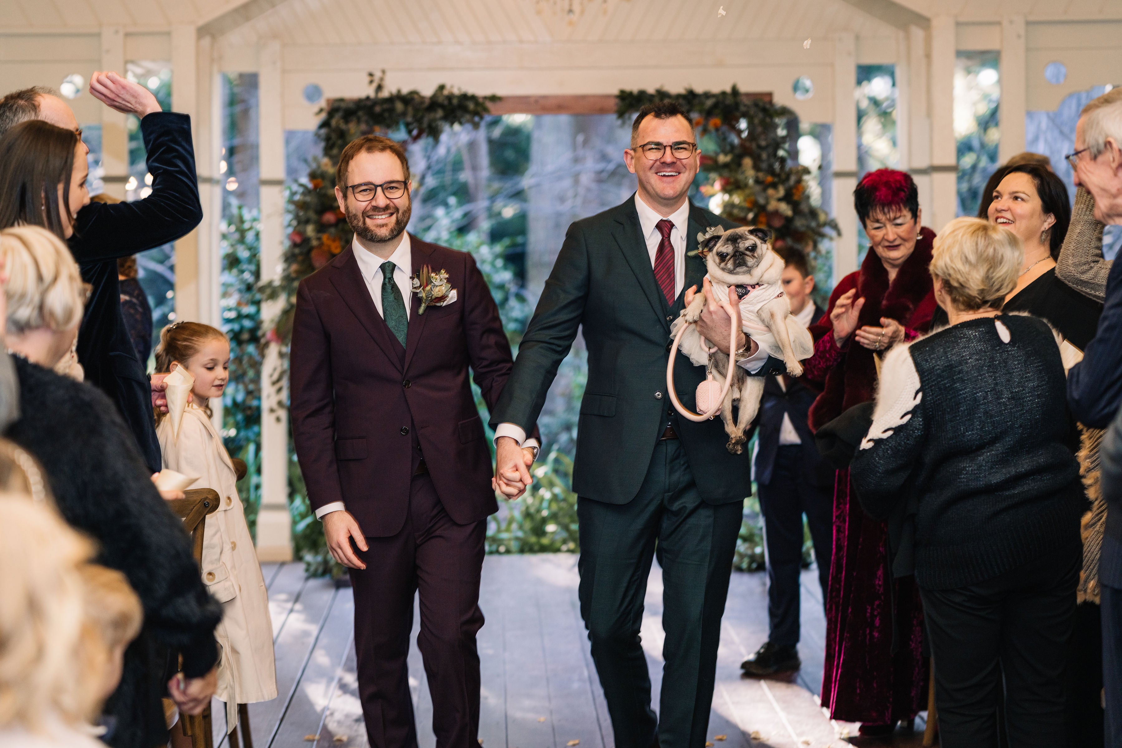 Two men walking down a aisle with a dog, surrounded by people in formal attire.