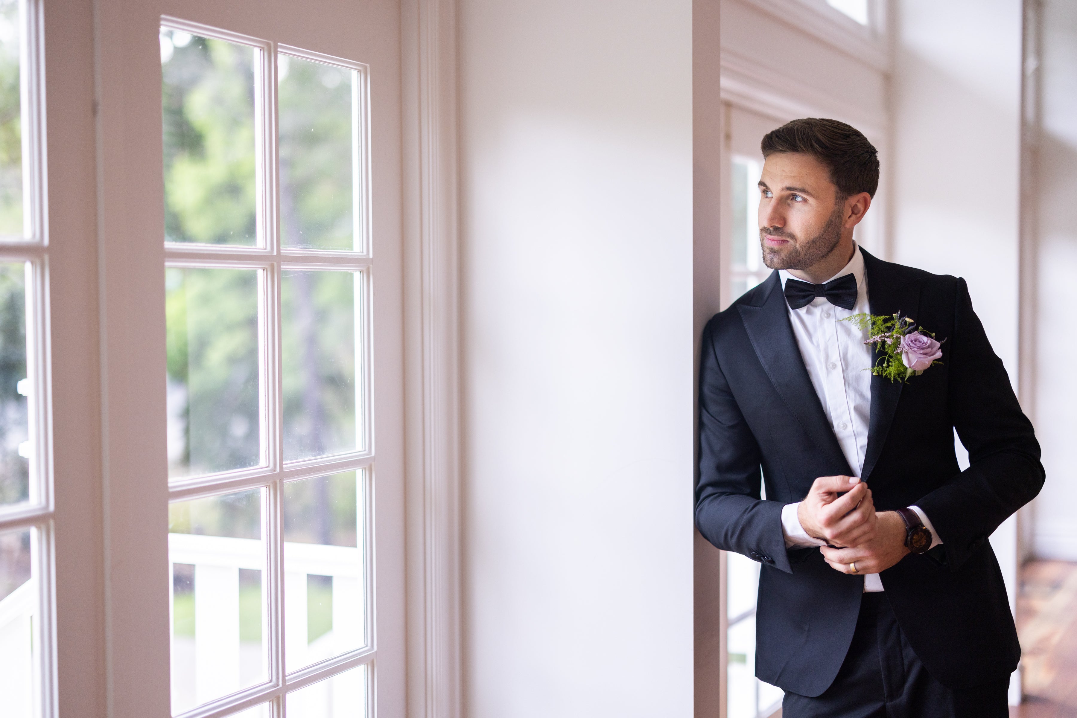 Man in a tuxedo standing by a window with a view of greenery