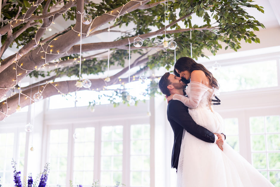 Couple in wedding attire embracing under a decorative tree with hanging lights and greenery.