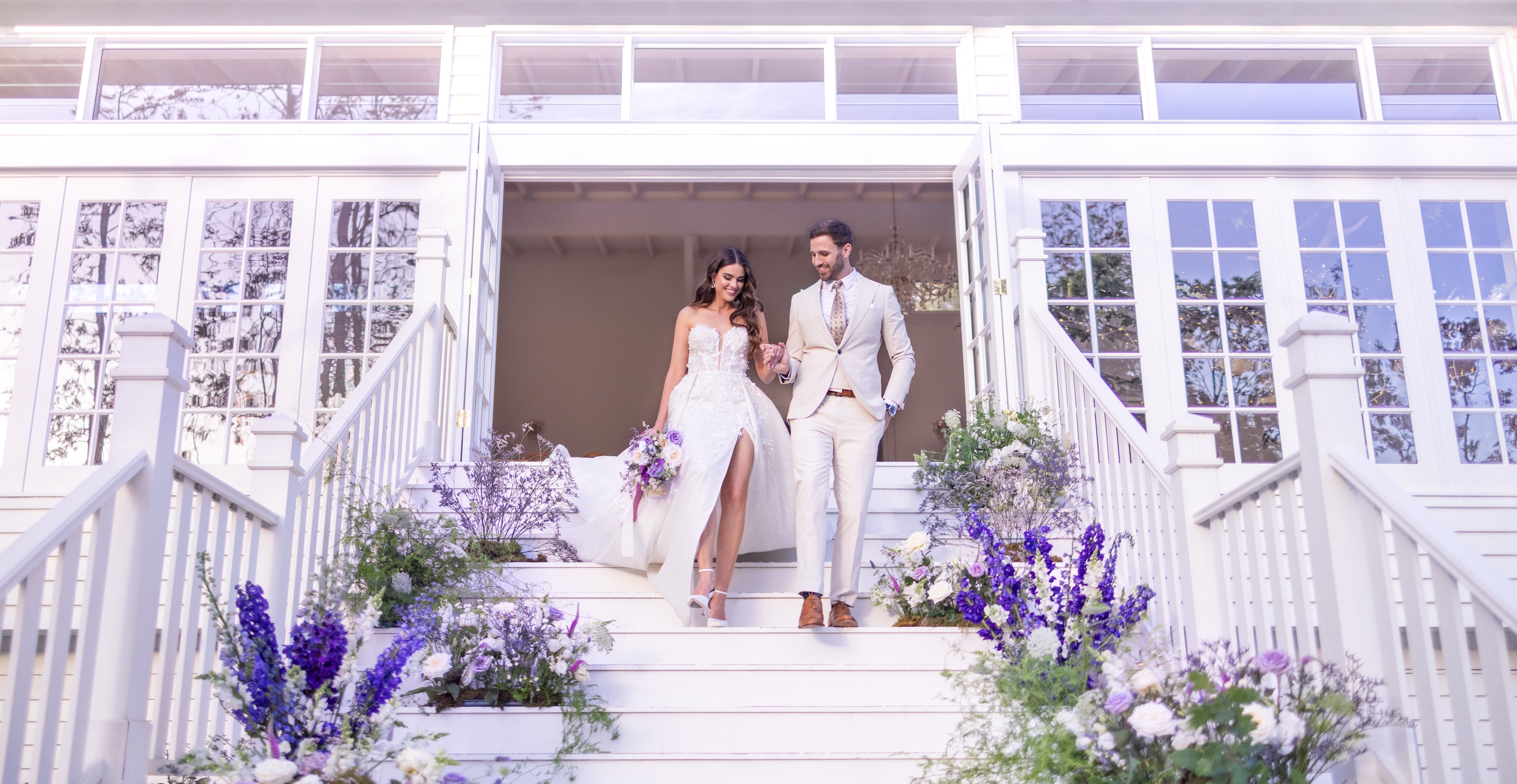 Couple standing on a white staircase with floral decorations, likely at a wedding venue.