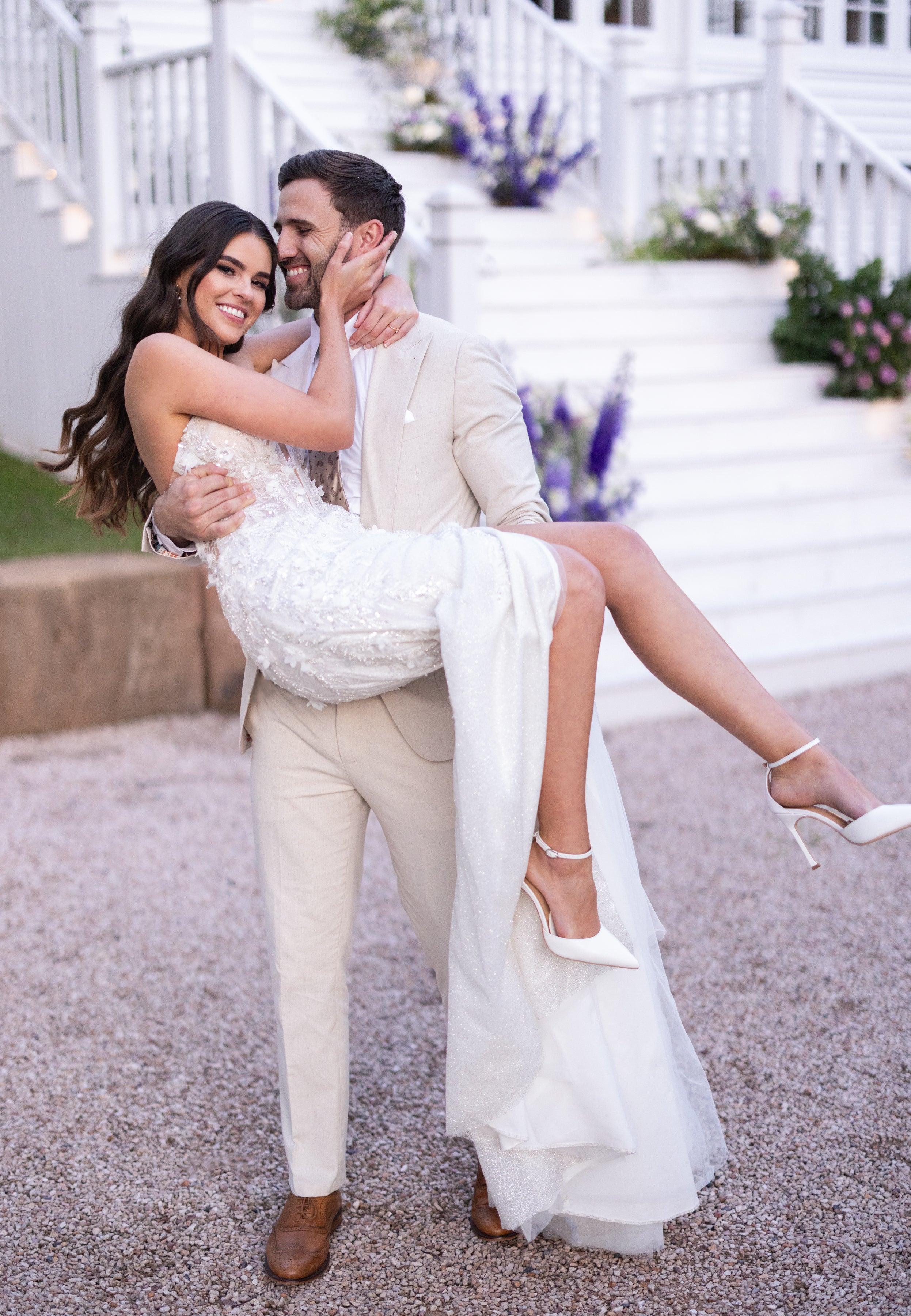 Man lifting a woman in a white dress on a gravel path with a white staircase and flowers in the background.