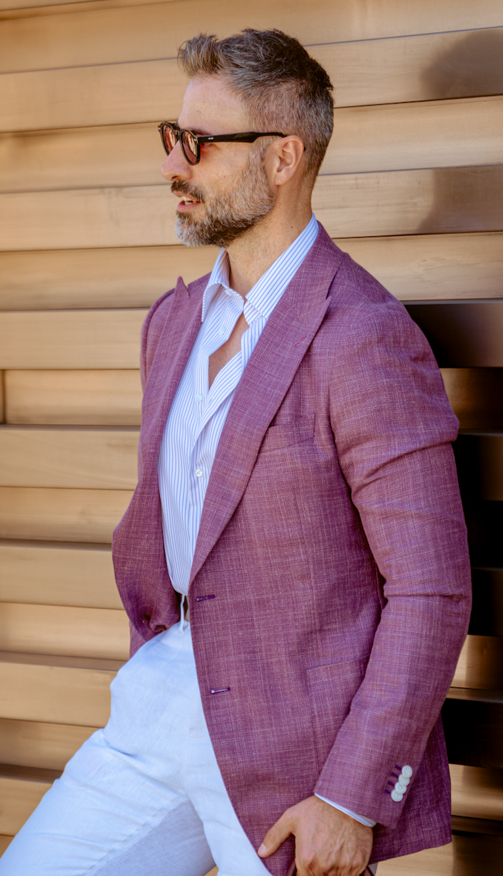 A man in a mauve-purple blazer, white shirt, and white trousers leans against a wooden slatted wall, holding a dark brown leather duffel bag.