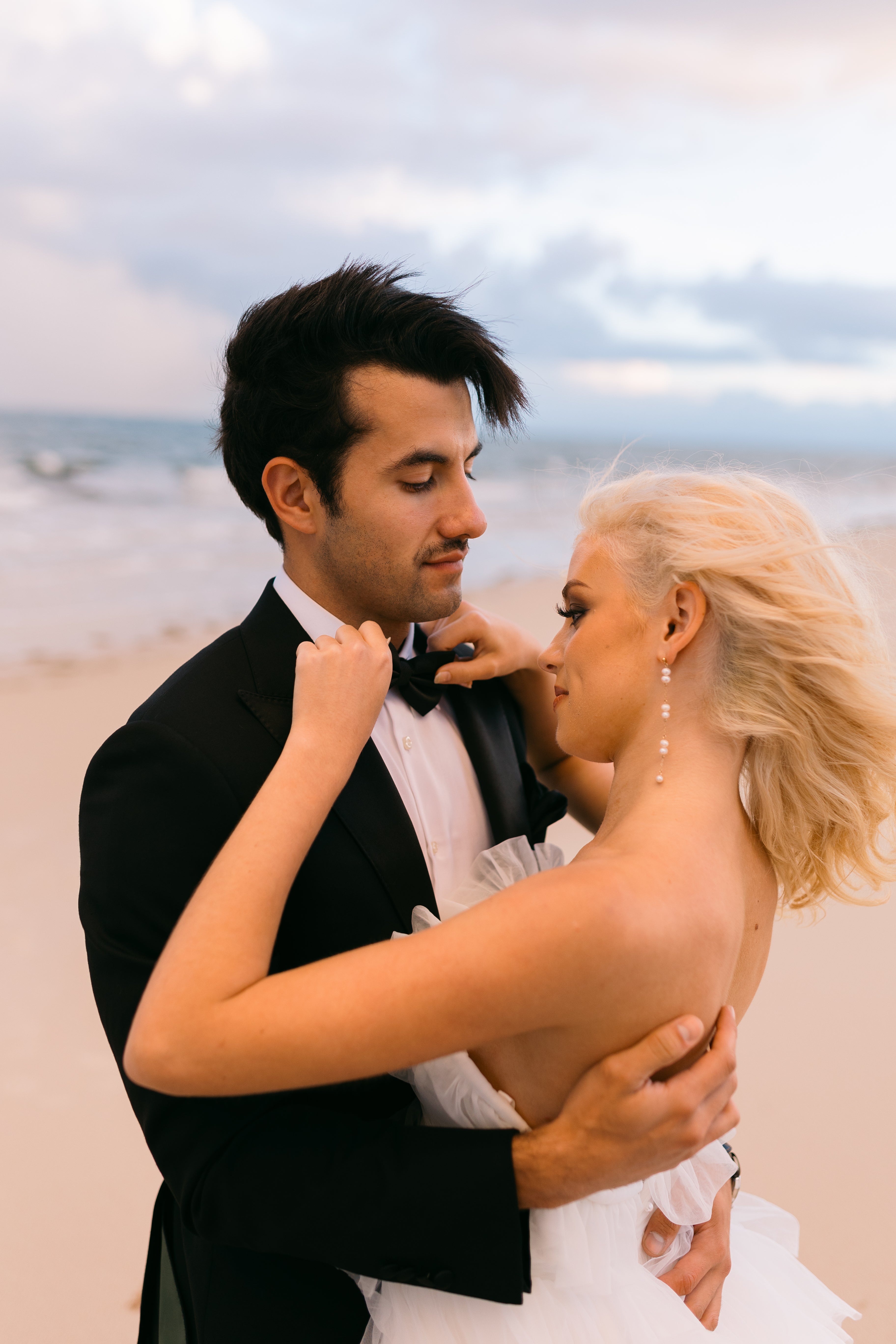 Man in a tuxedo and woman in a white dress embracing on a beach.