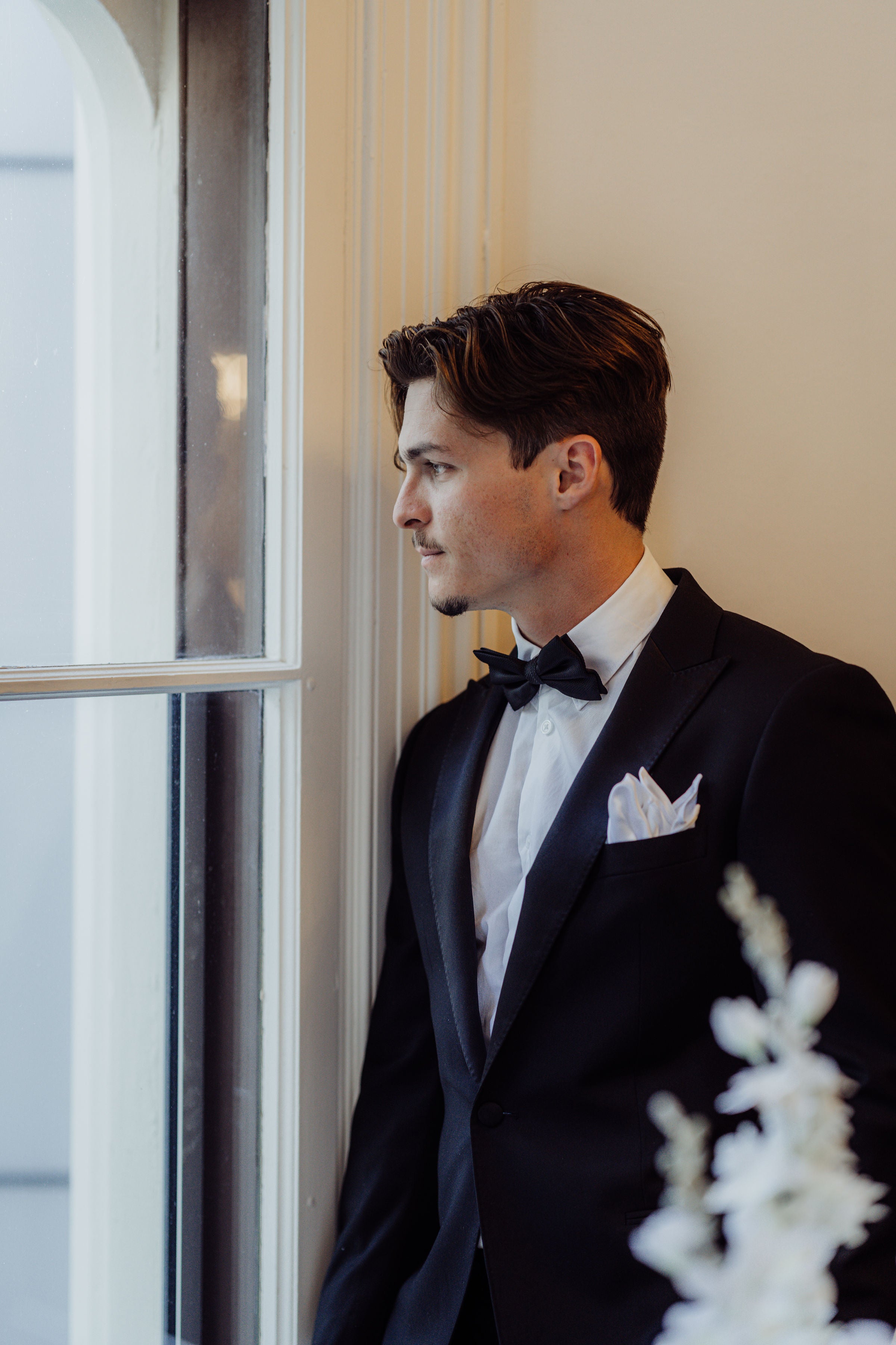 Man in a black tuxedo standing by a window with a neutral background