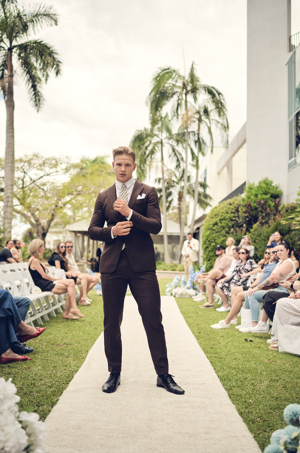 Man in a suit standing on a wedding aisle with guests and palm trees in the background