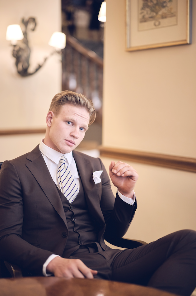 Man in a formal suit sitting in an elegant room with a painting on the wall.