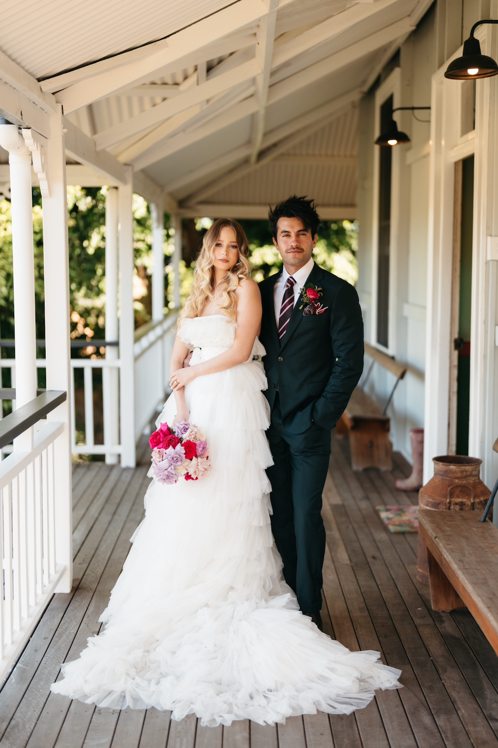 Wedding couple standing on a wooden porch with a white house in the background.