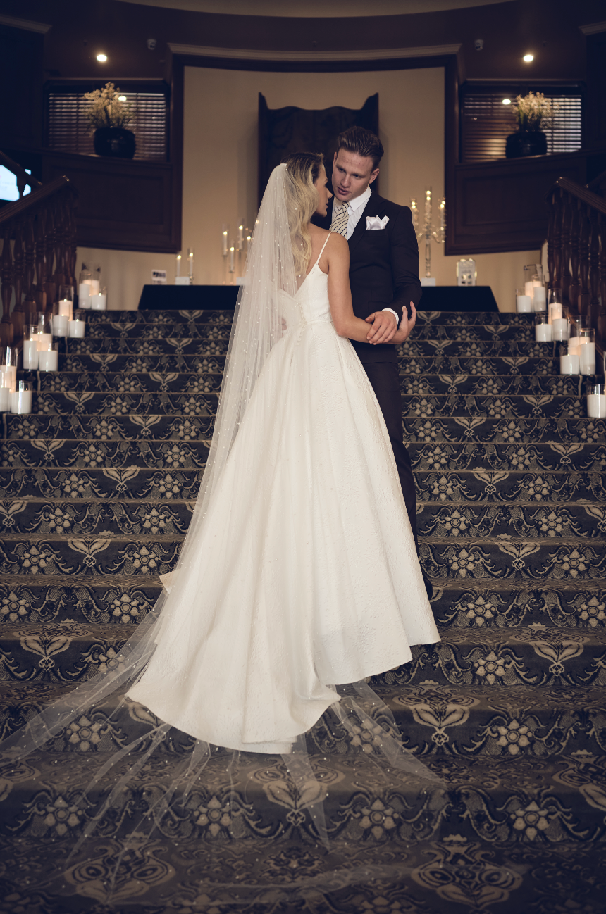 Couple in wedding attire standing on a grand staircase with candles and decorative lights.