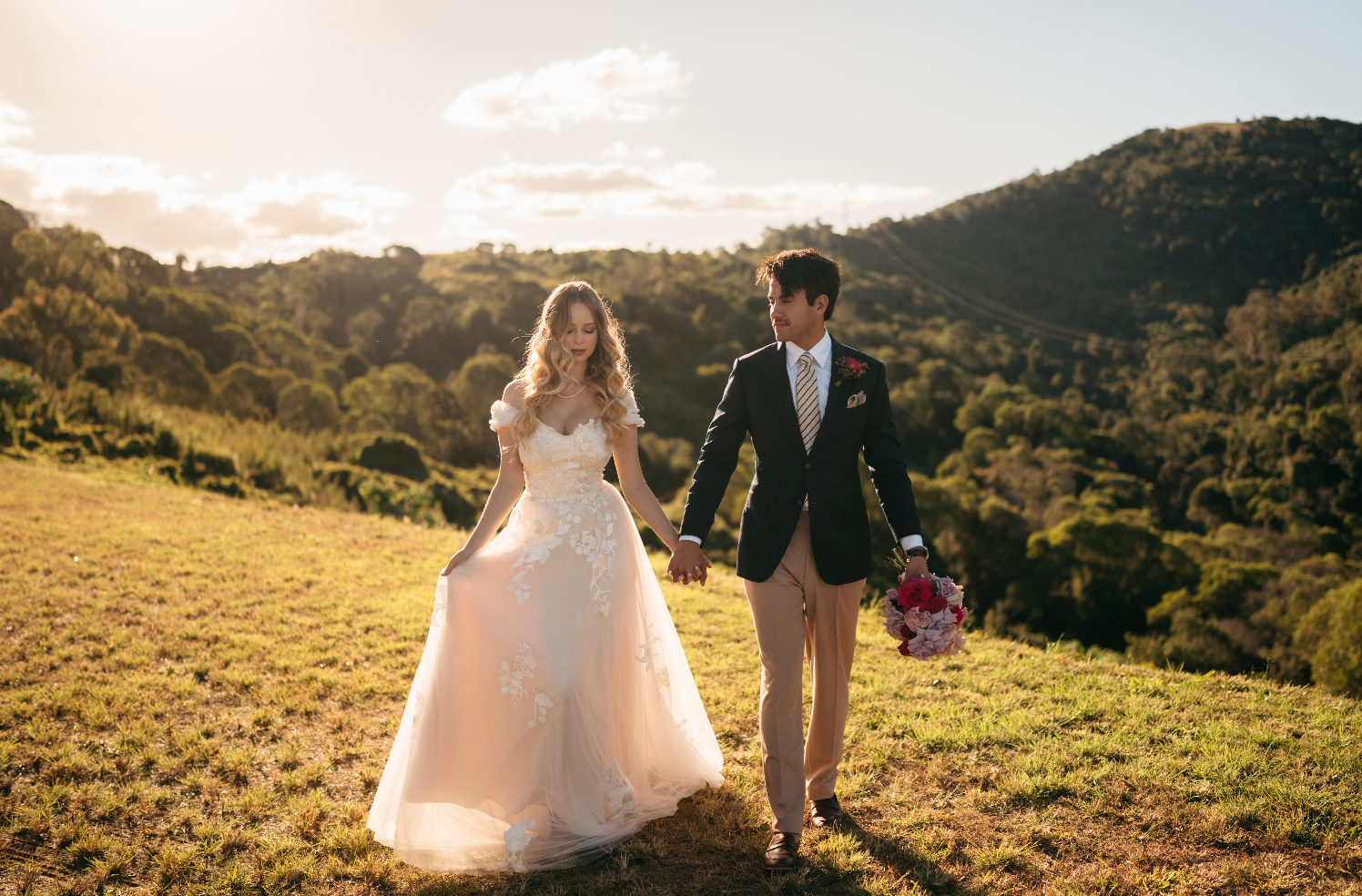 Couple in wedding attire walking hand in hand on a grassy hill with a scenic background.