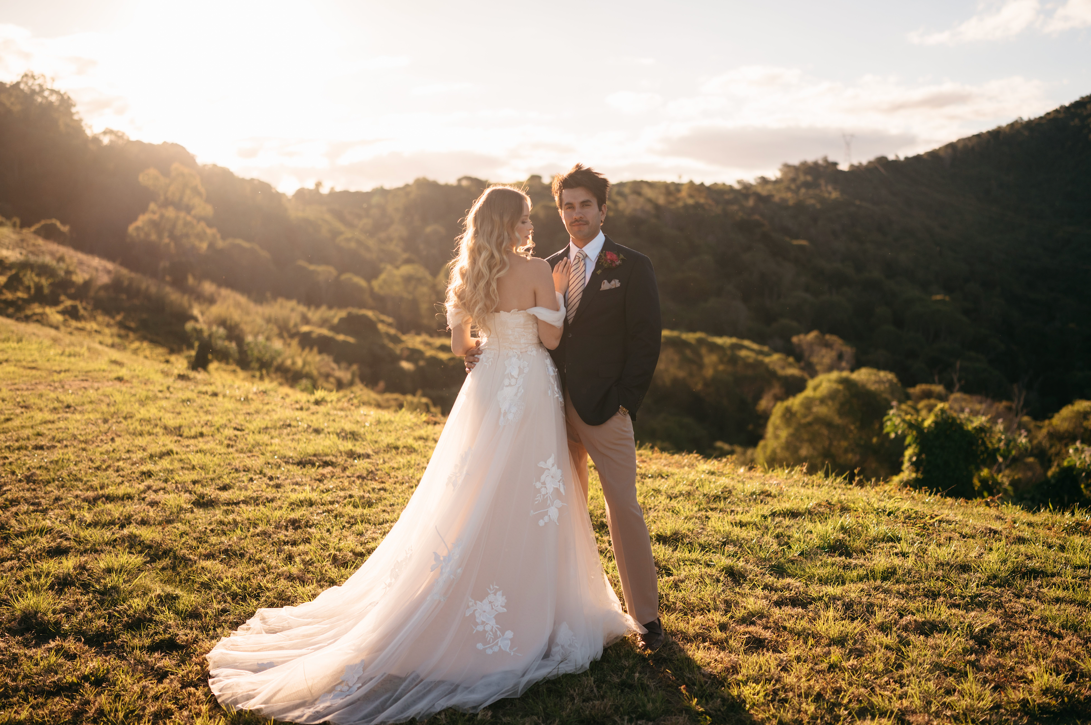 Couple in wedding attire standing in a scenic outdoor setting with mountains in the background.