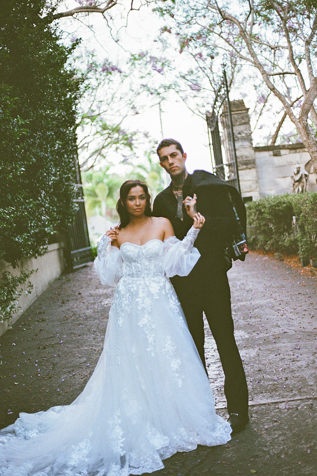 Man and woman in formal attire standing on a path with trees and a building in the background