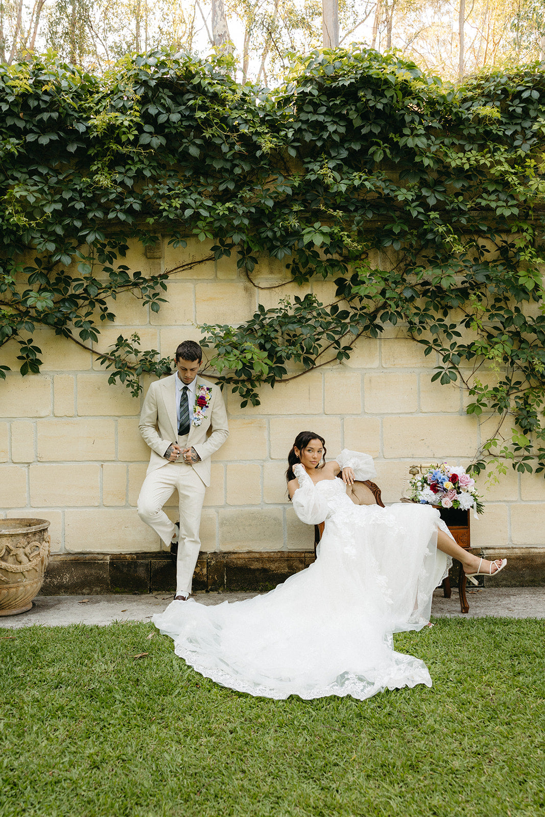Couple in wedding attire sitting on a bench with greenery and a stone wall in the background