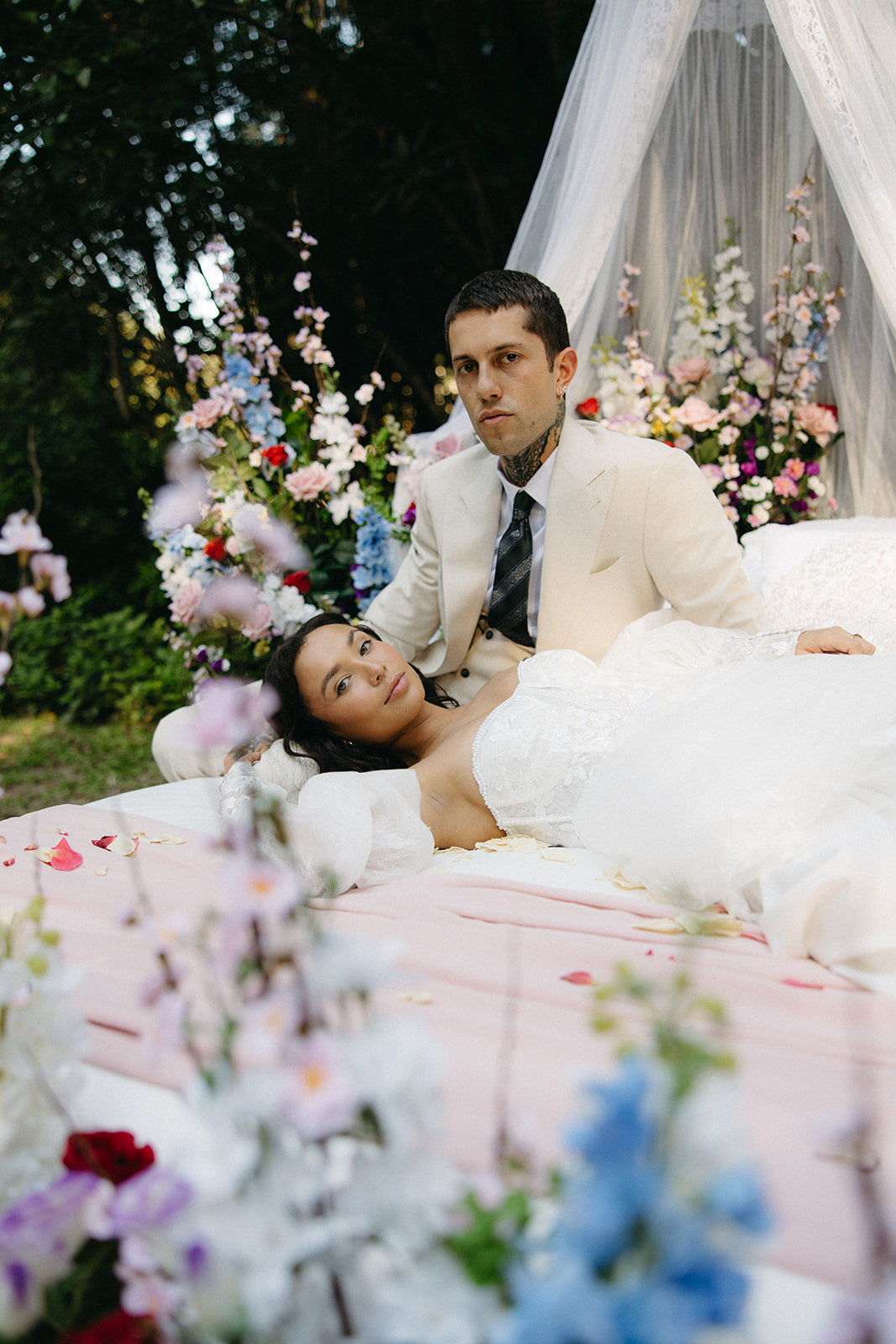 Couple lying on a bed surrounded by flowers and a canopy in an outdoor setting