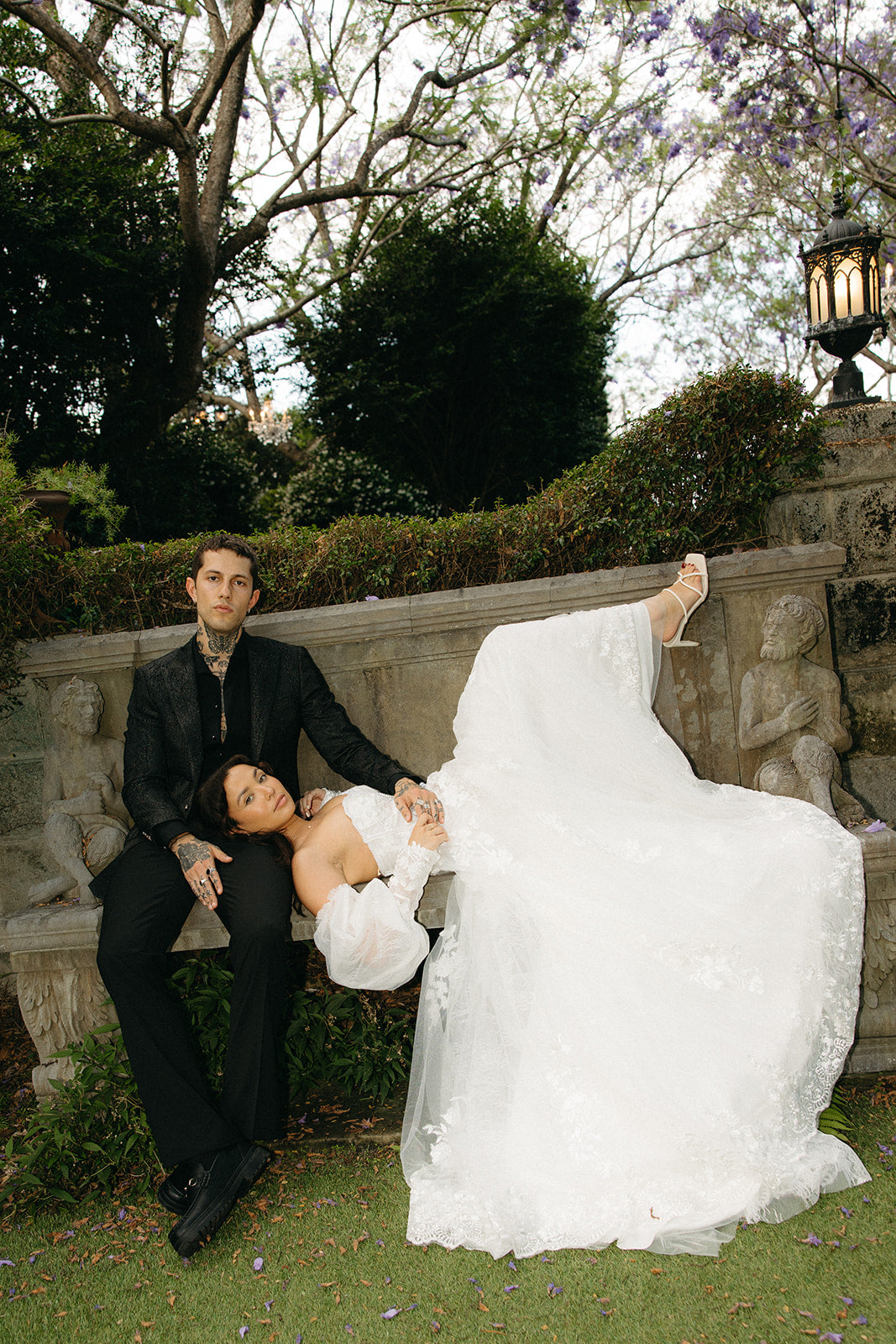 Man and woman in wedding attire sitting on a stone bench outdoors.