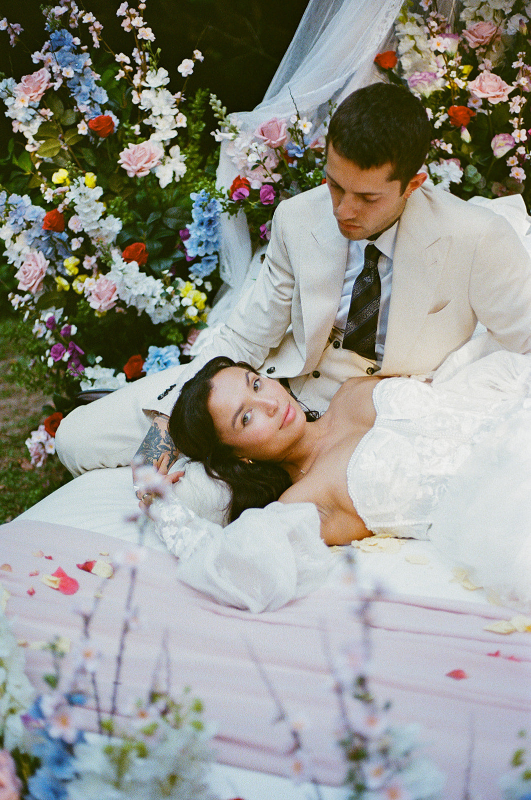 Couple in wedding attire lying on a bed of flowers with floral decorations in the background